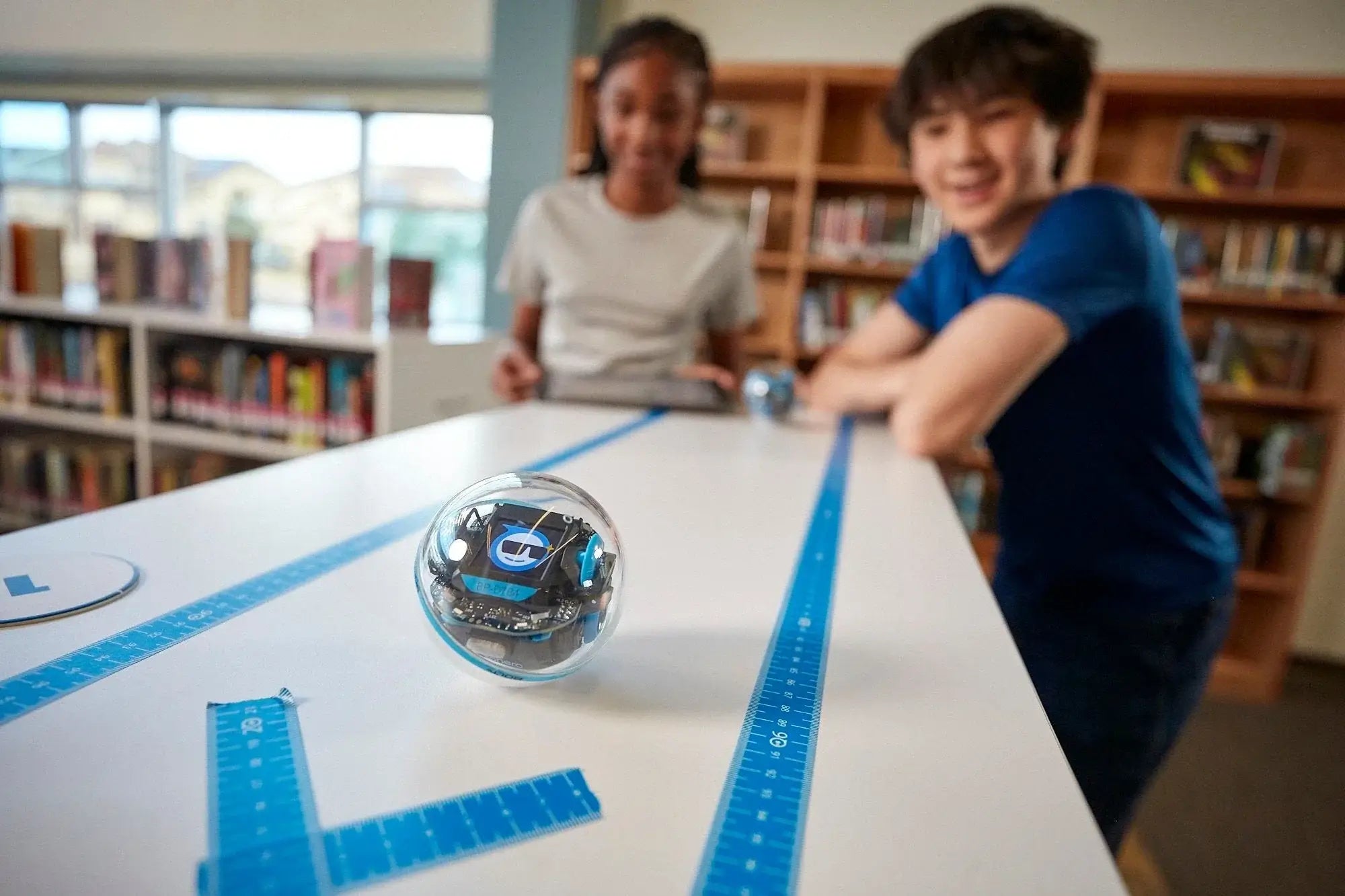 Two students engaging with a Sphero robot on a table in a library, promoting STEM learning and robotics education.