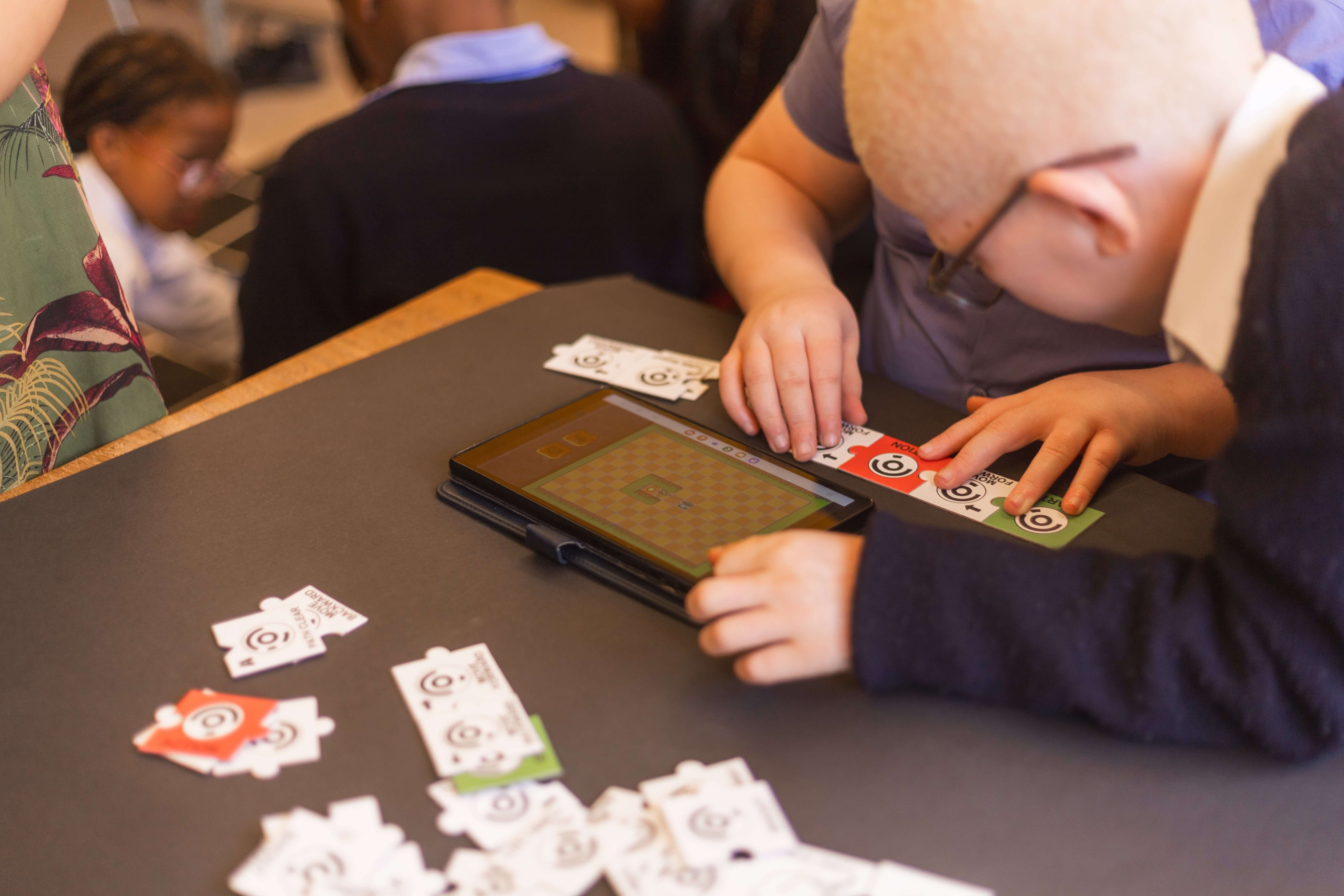 Children playing a card game on a tablet at school, engaging with colorful cards on a table.
