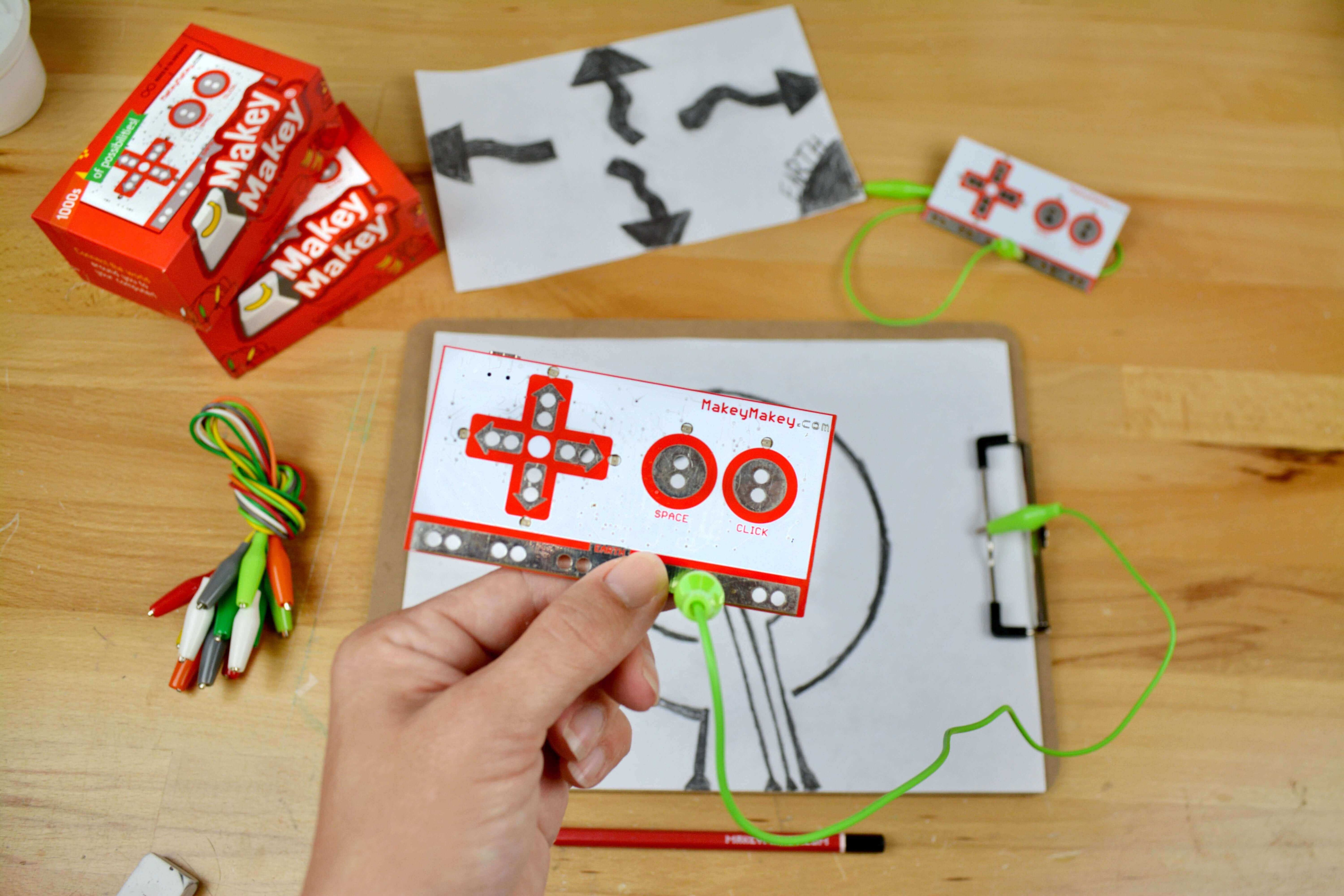 Person holding a Makey Makey board with colored pencils and sketches on a wooden table.