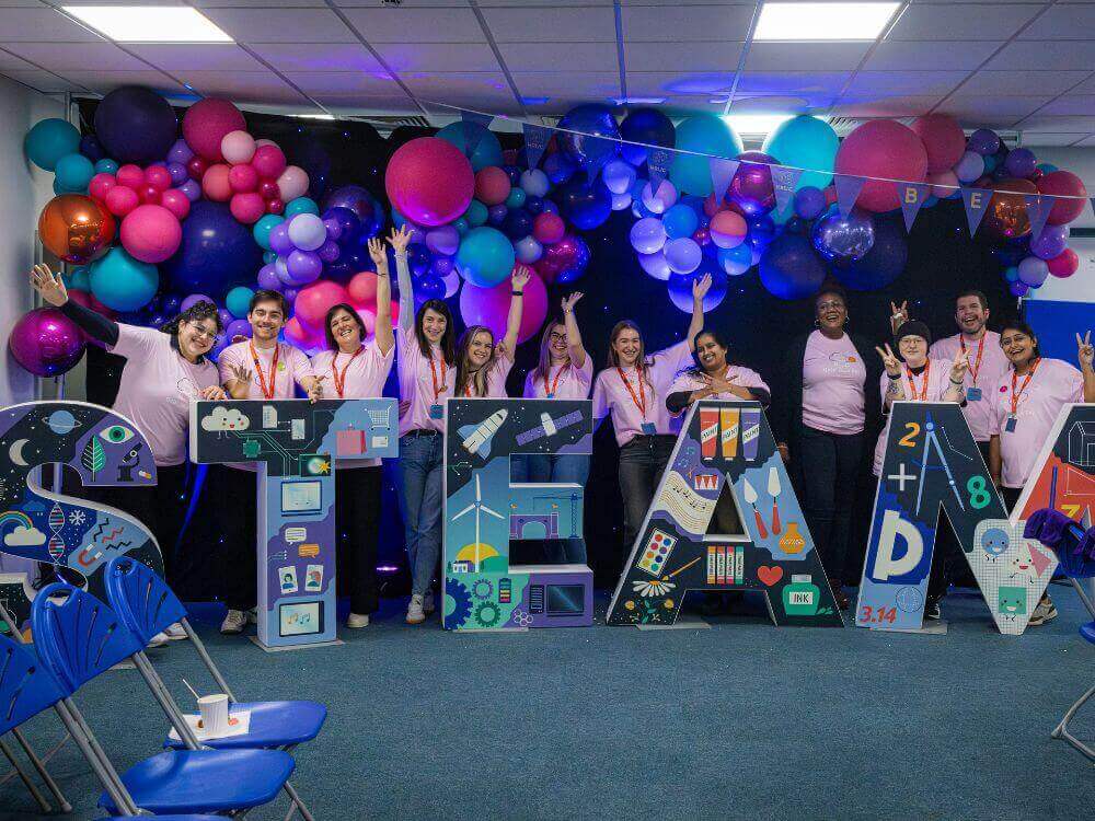 A group of smiling individuals in pink shirts celebrate STEAM education with colorful balloons in the background.