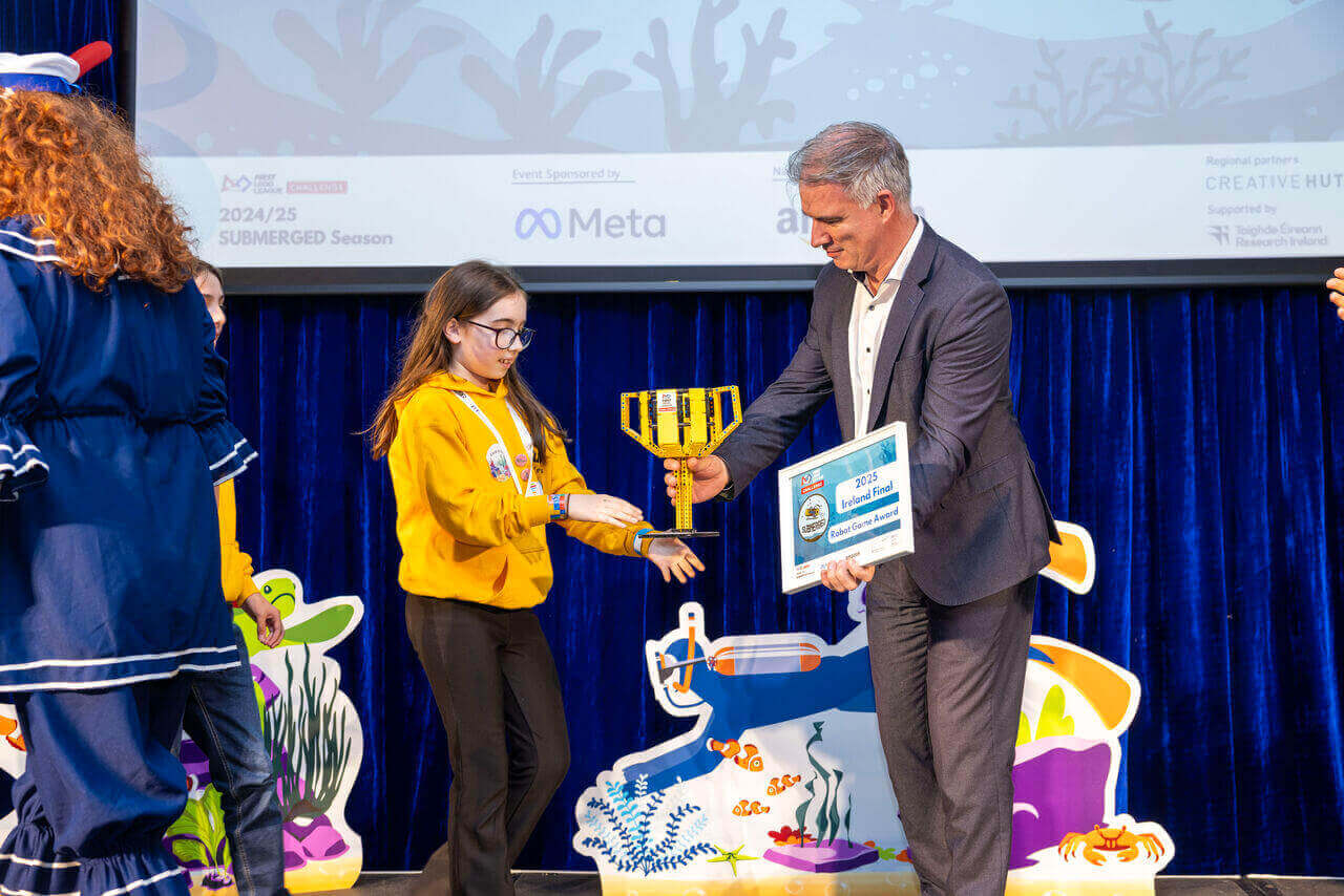 Child receiving an award from an adult at a ceremony, with colorful decorations in the background.