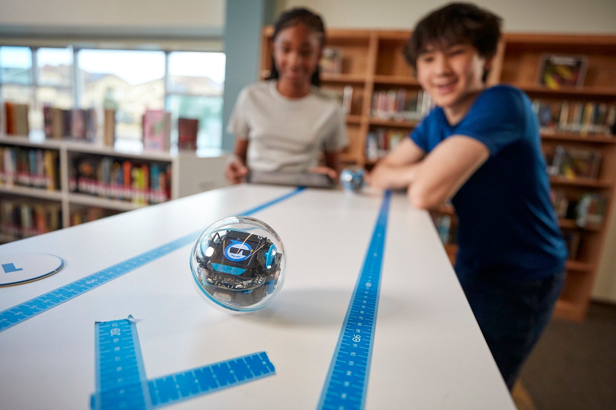 Two students engaging with a robot on a table in a library, surrounded by rulers and learning materials.