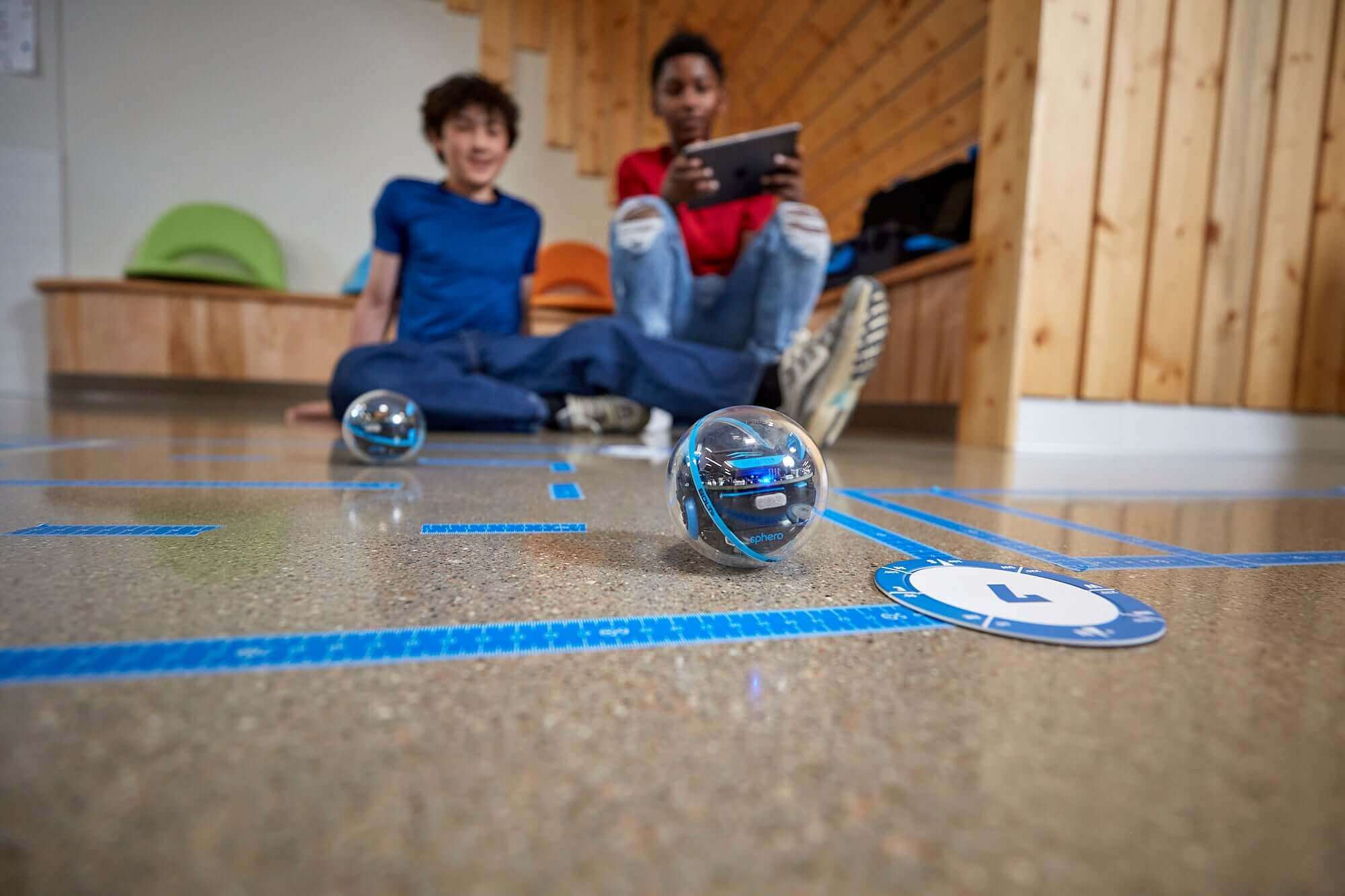 Two students engaging with Sphero robots on a coding mat while using a tablet for interactive learning.