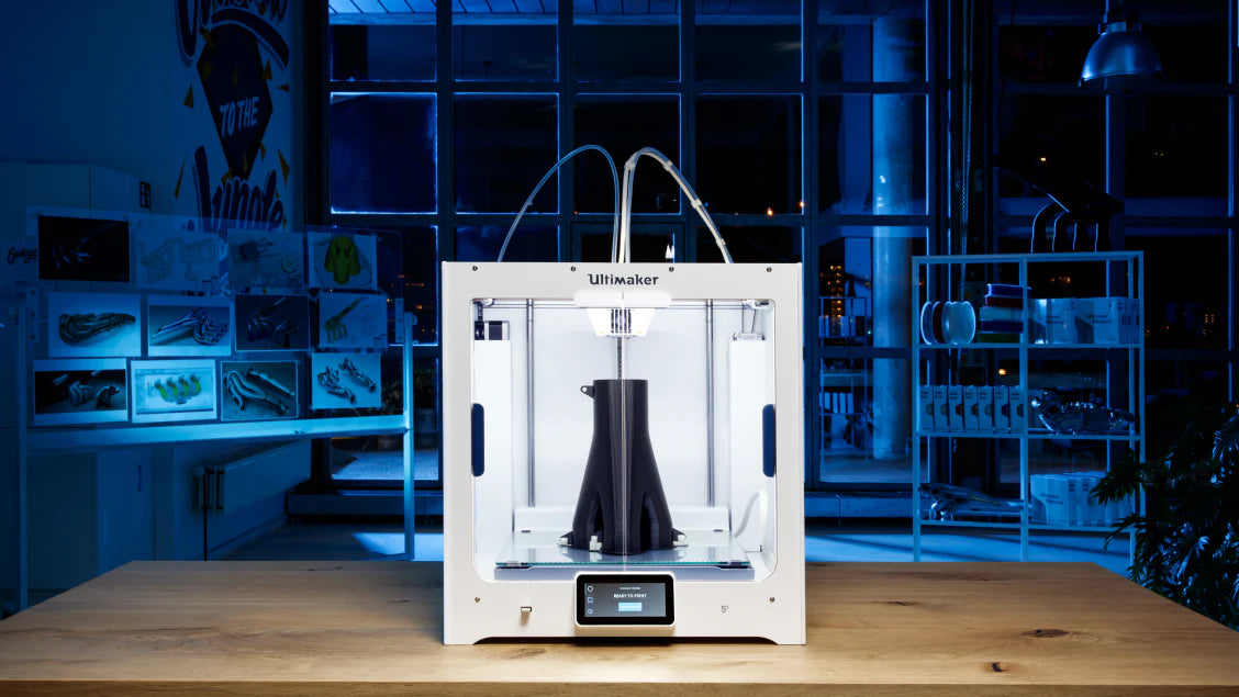 Ultimaker 3D printer in a modern workspace, creating a black object under blue lighting.