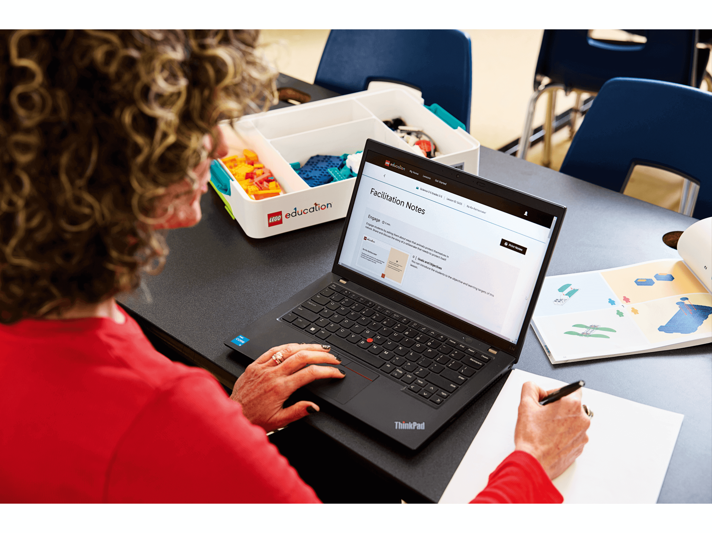 Woman using a laptop for educational facilitation notes, surrounded by colorful learning materials in a classroom.
