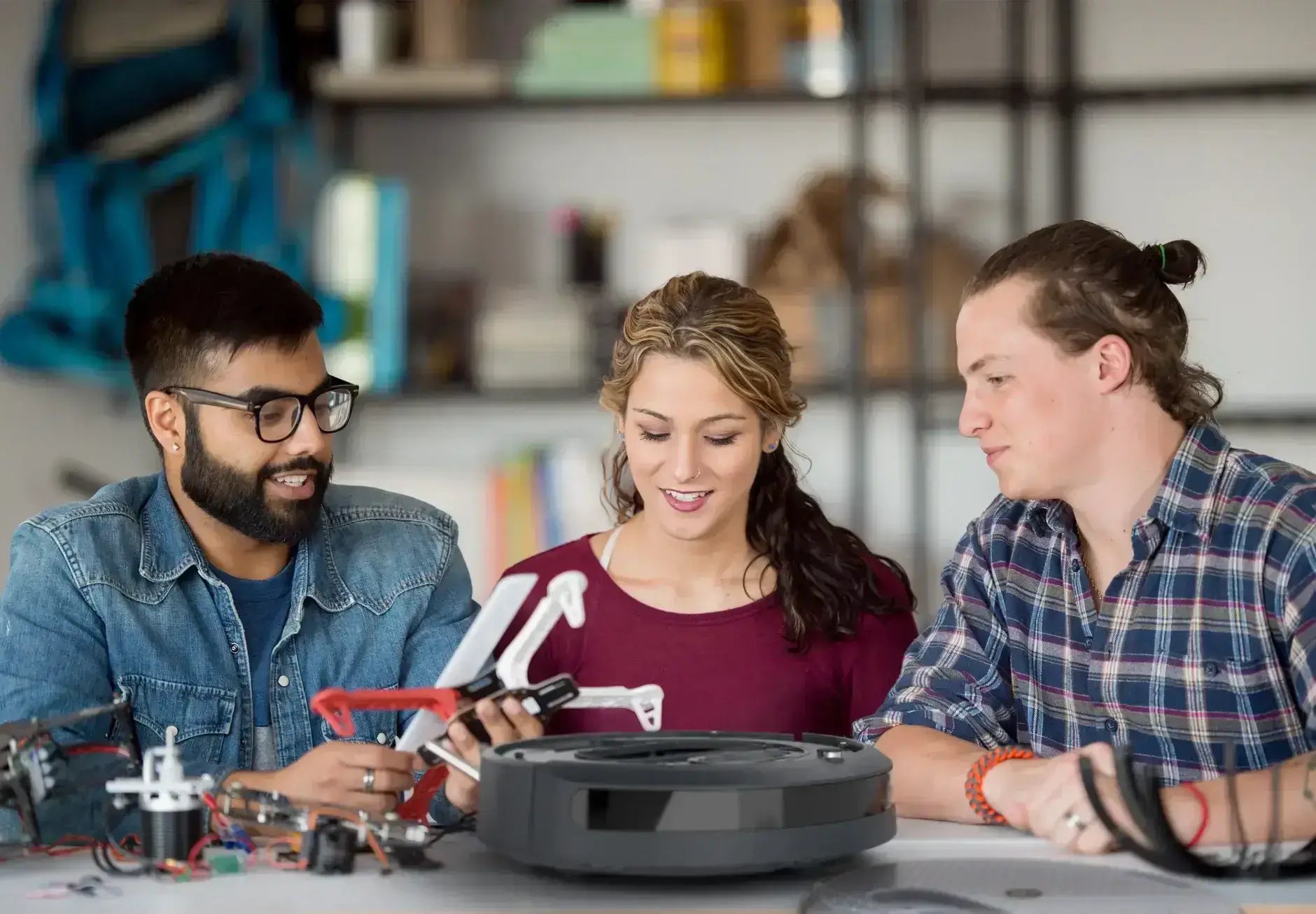 Three individuals working on tech gadgets, including a drone and a robotic vacuum, in a collaborative workspace.