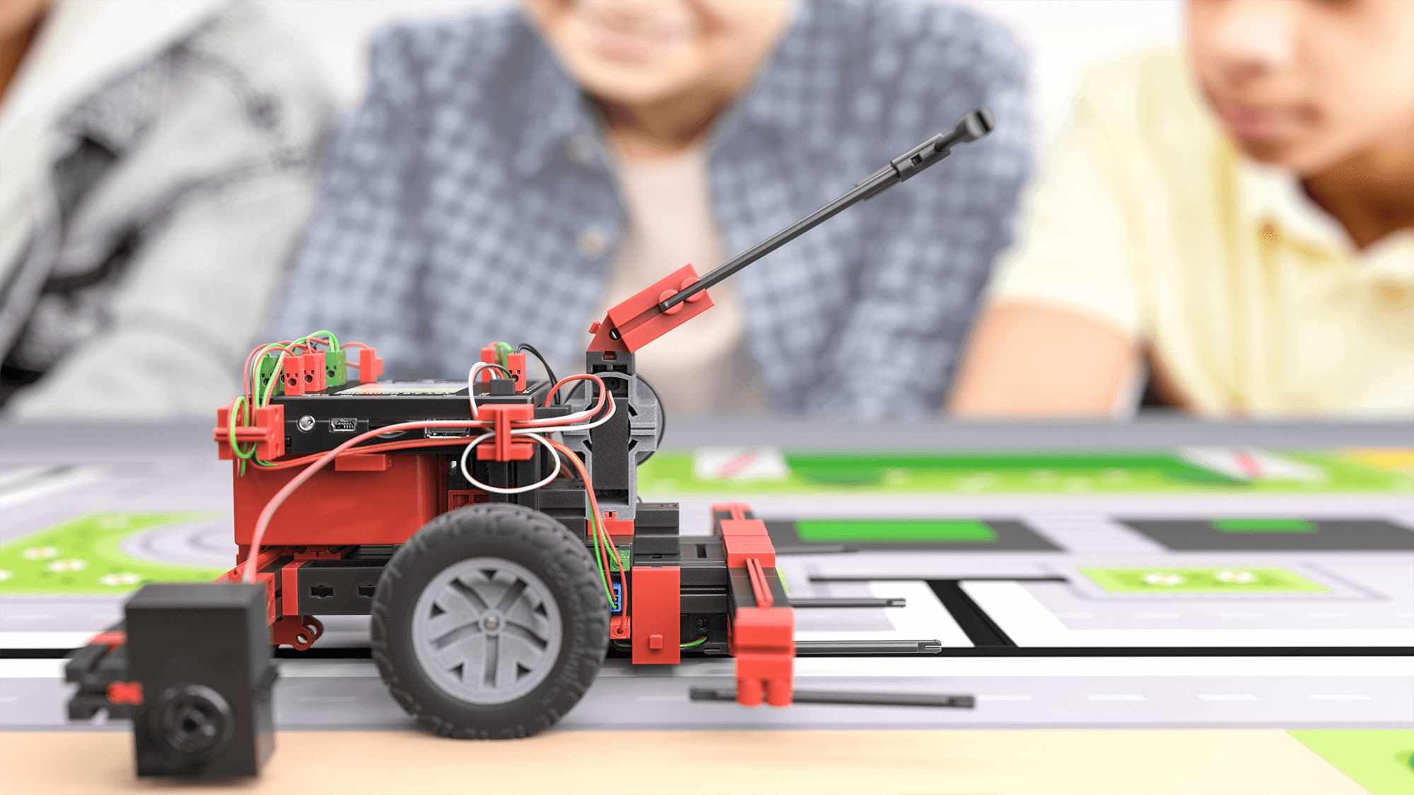 A close-up of a robotic vehicle with wires, focused on coding and robotics education. Kids are engaged in the background.