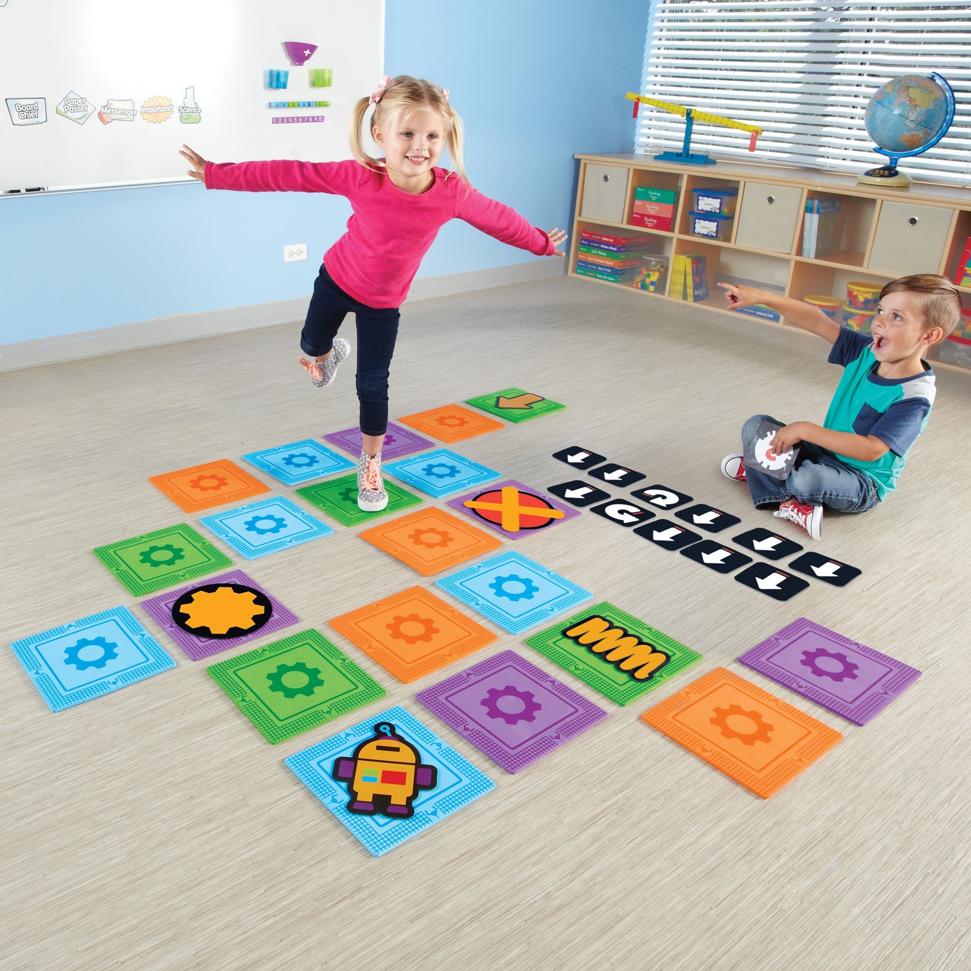 Two children playing with a life-size coding maze. One child balances on a foam mat while the other points and holds a coding card, demonstrating active, screen-free programming.