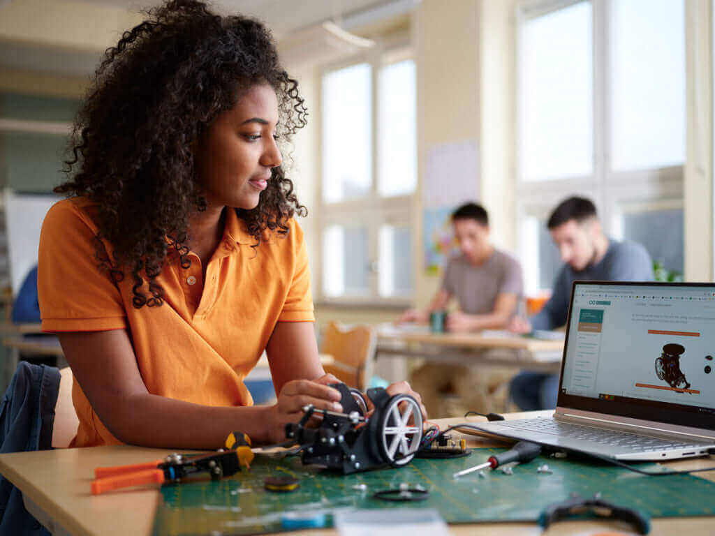 Young woman assembling a robot in a workshop, focused on her laptop with classmates in the background.