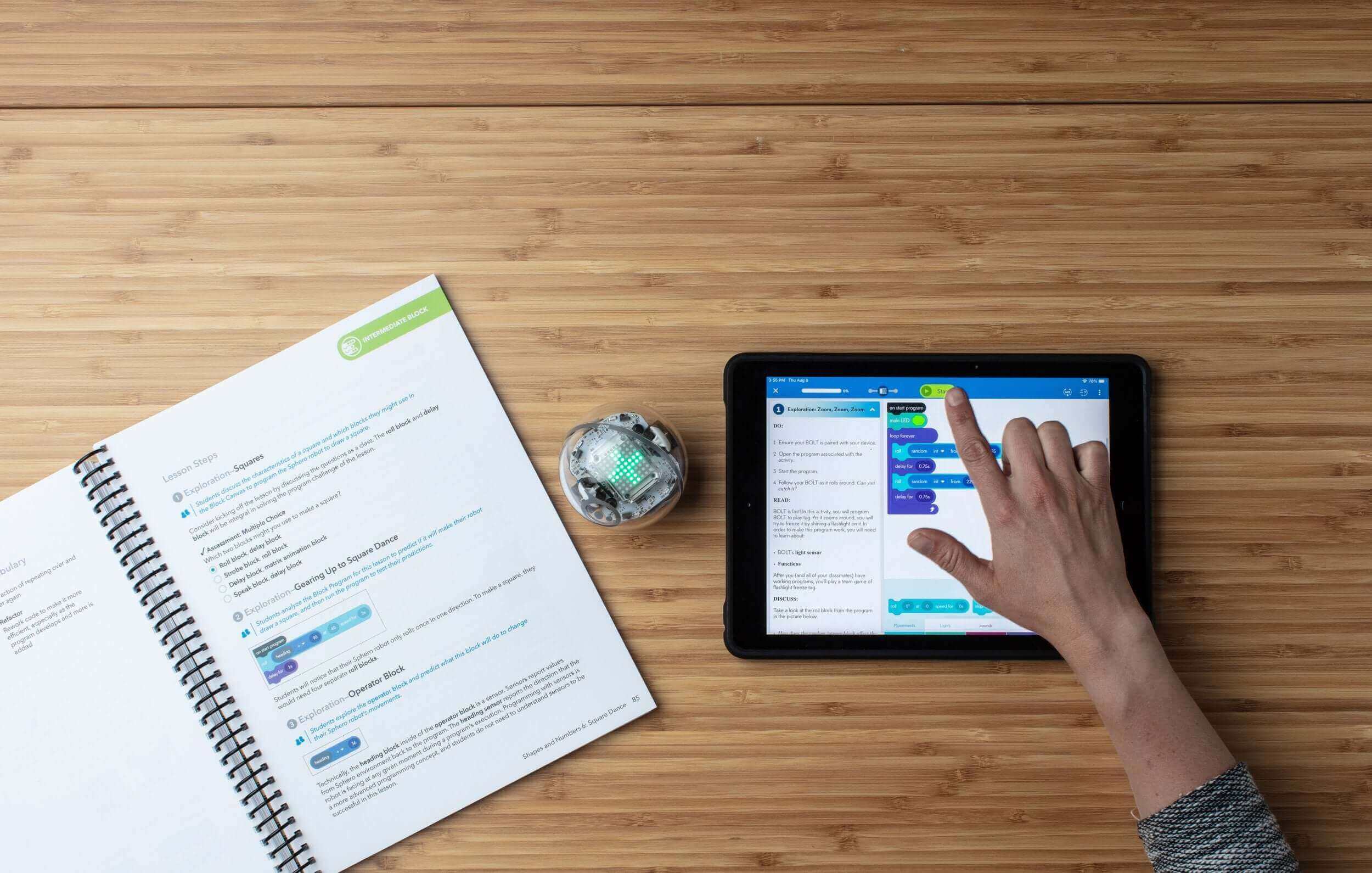 Person programming a robot using a tablet and an educational book on a wooden table.