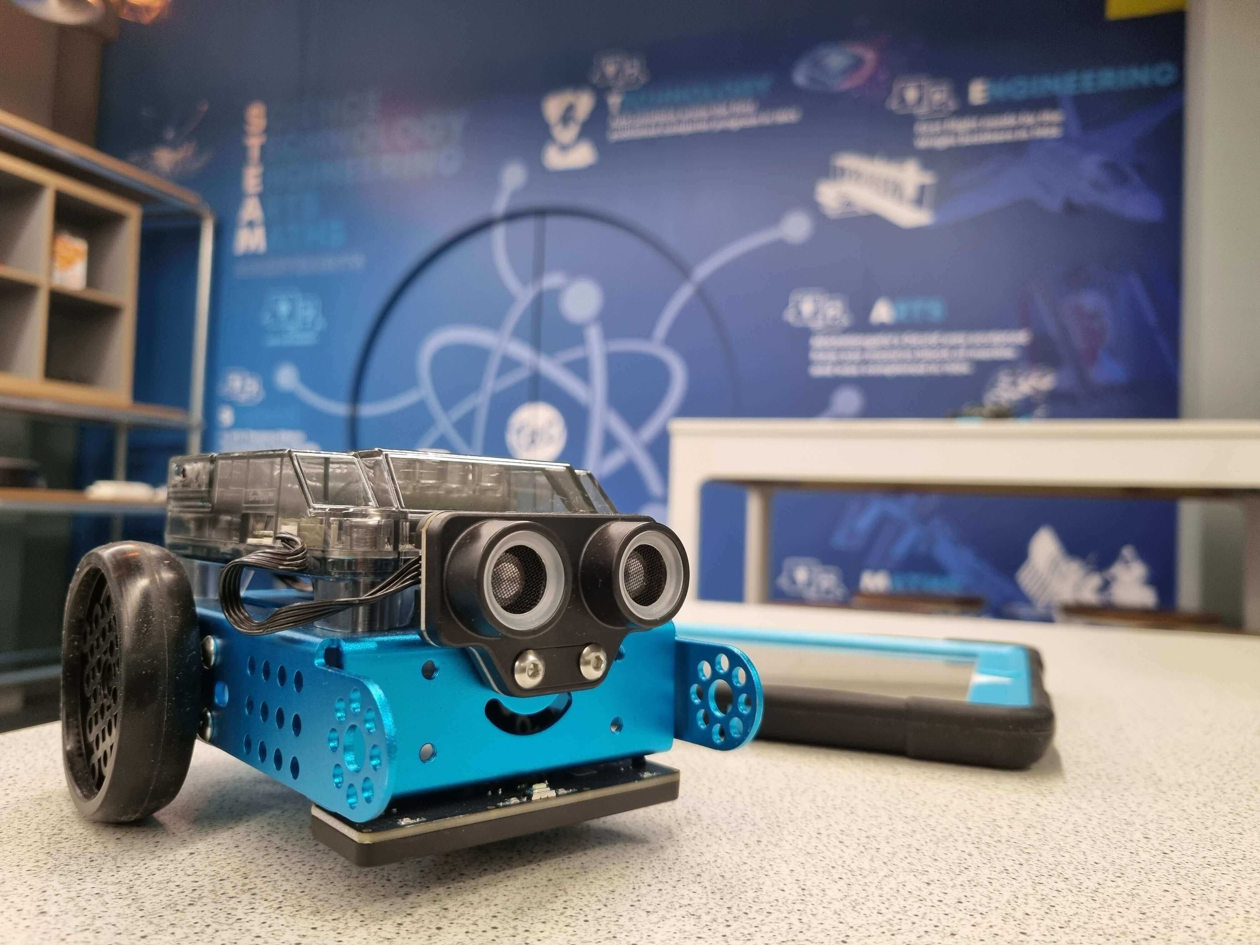 A playful blue robot with wheels and smiling face, displayed in a tech-themed educational environment.