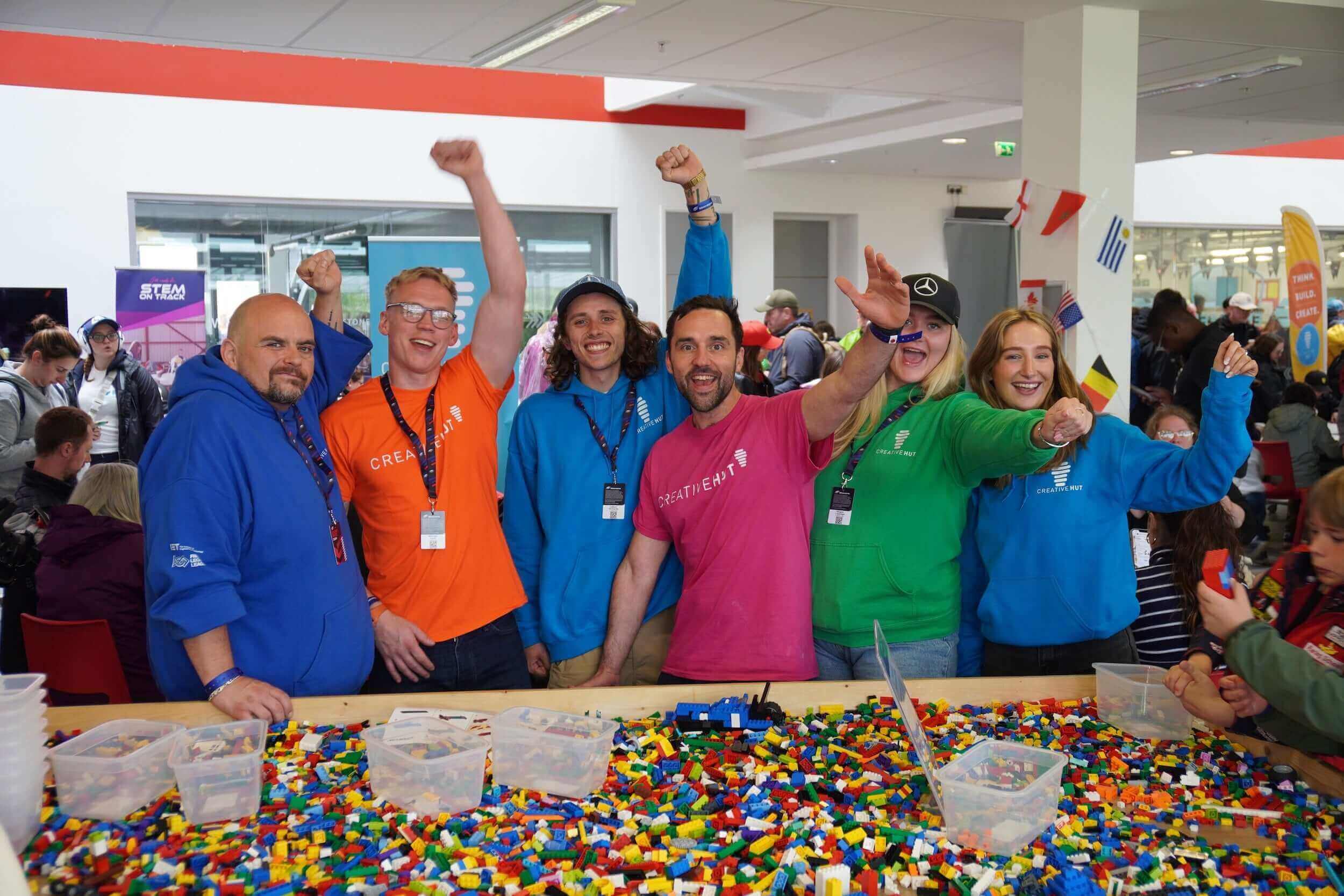 A group of enthusiastic organizers wearing colorful hoodies celebrates at a LEGO building event.