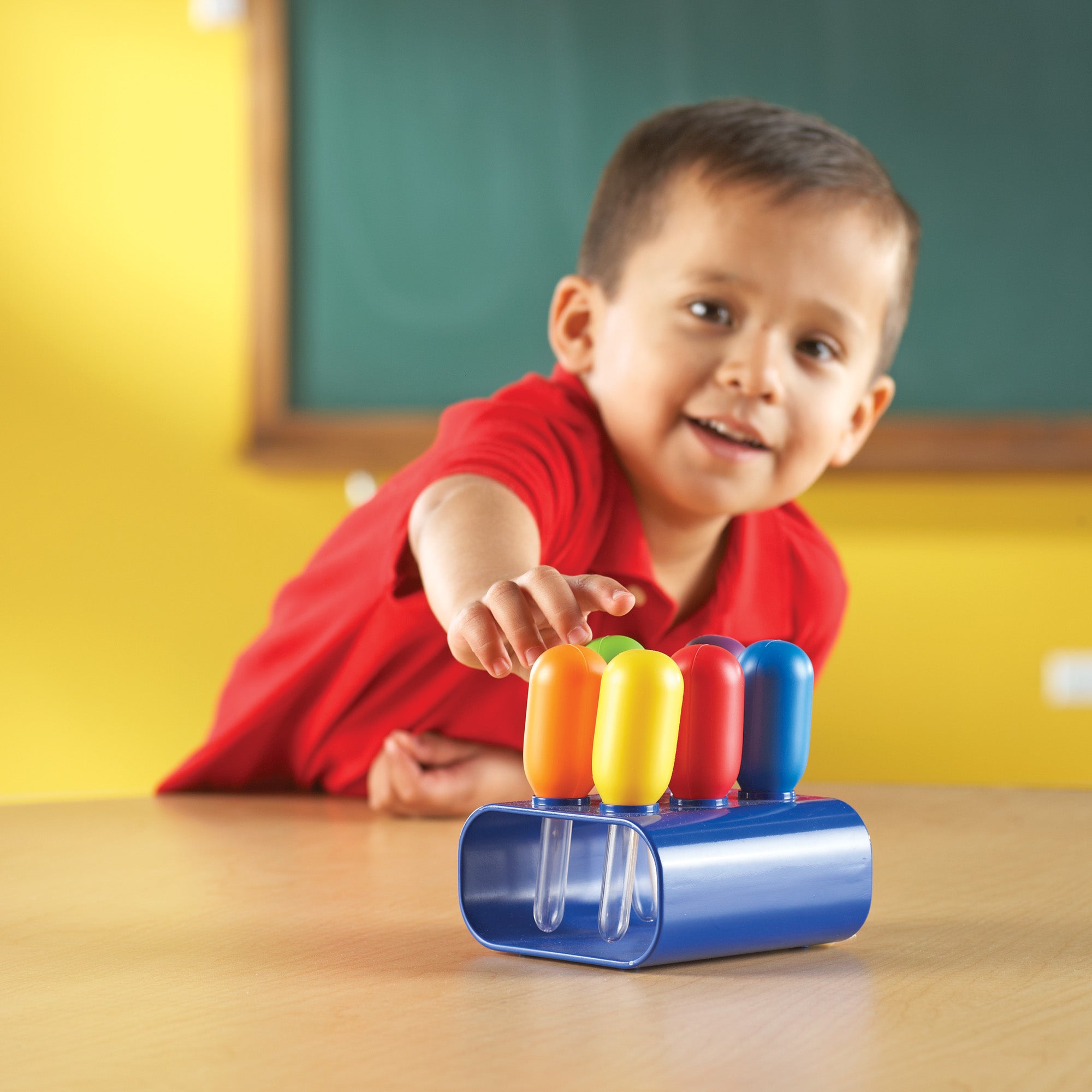 A young child in a red shirt smiles while reaching for a set of colourful Jumbo Eyedroppers in a classroom setting. The droppers, featuring chunky bulbs in orange, yellow, red, and blue, are neatly stored upright in a sturdy blue plastic stand on a wooden table. A chalkboard is visible in the soft-focus background. The image highlights the oversized, child-friendly design of the 11.5cm droppers, demonstrating their use for sensory play and early scientific exploration for children aged 3 and up.