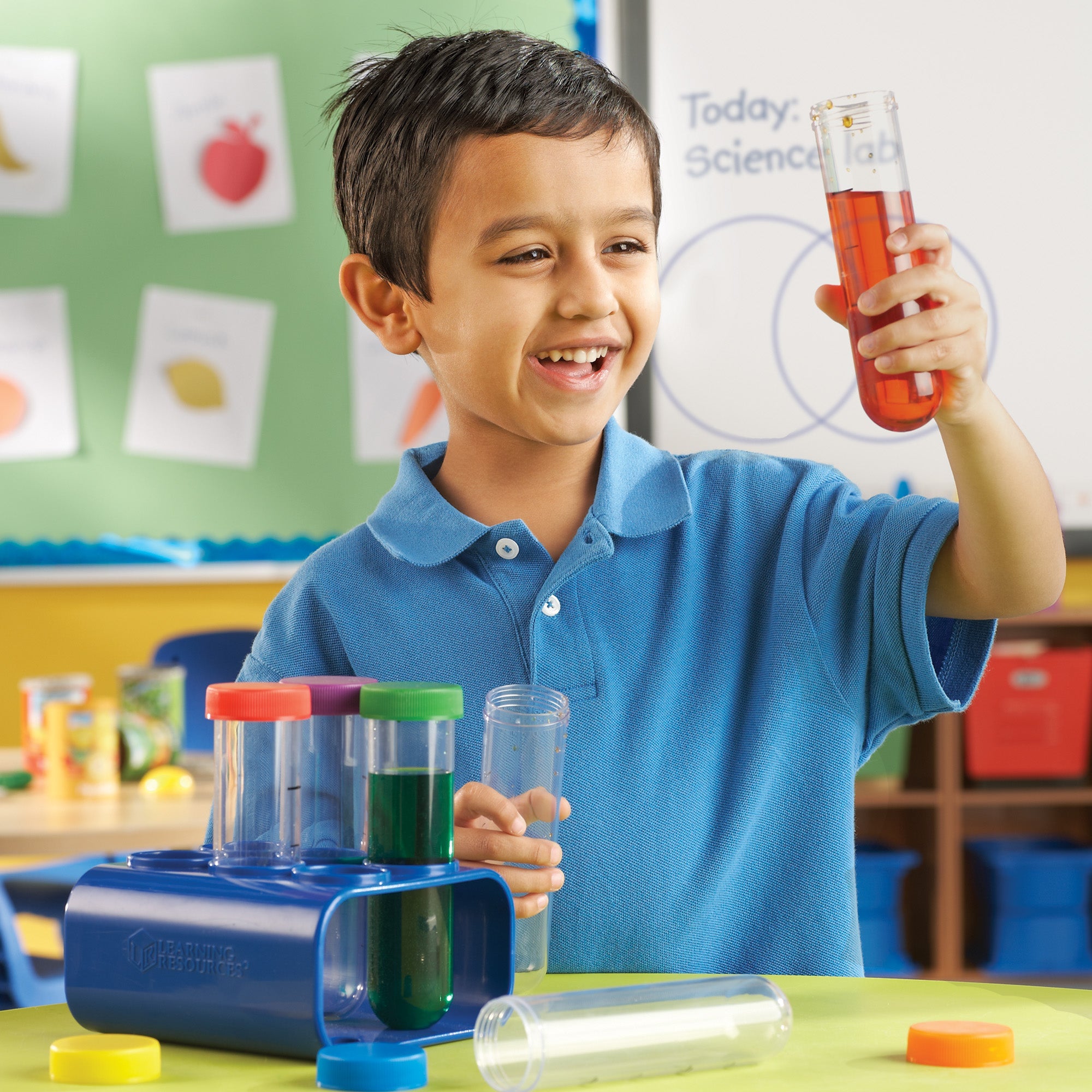 A young boy in a blue polo shirt smiles enthusiastically while holding up a Primary Science® Jumbo Test Tube filled with orange liquid. He is in a brightly lit classroom with a "Science Lab" sign on a whiteboard behind him. On the table sits the blue storage stand holding several other 15cm test tubes, including one filled with green liquid. The image showcases the durable, child-friendly design and the colourful screw-on lids, illustrating hands-on STEM learning and scientific observation for children aged
