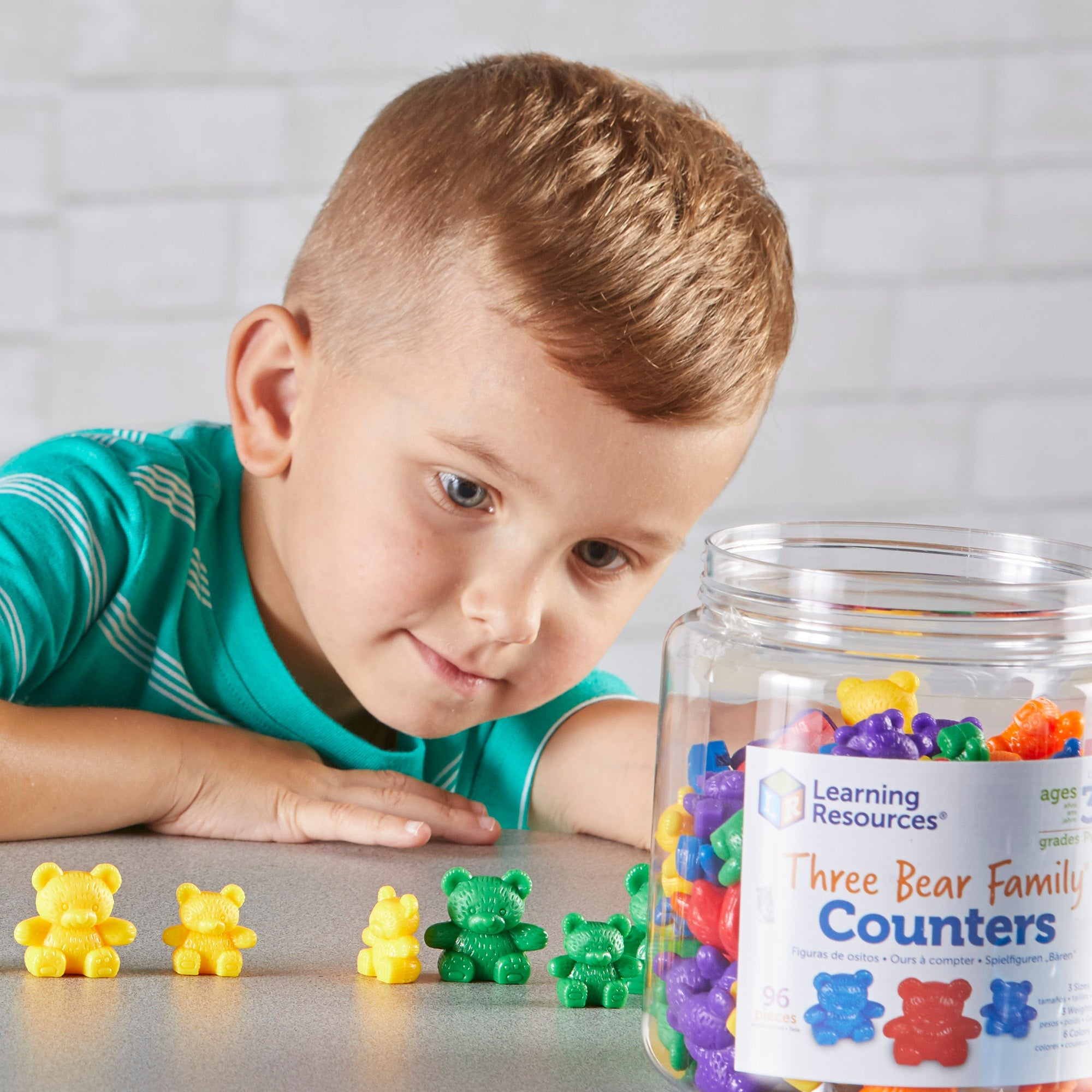 A young boy leans over a table, intently focused on sorting and lining up colourful Three Bear Family counters. In front of him, he has arranged several yellow and green bears by size. To the right, a large, clear storage jar labelled "Learning Resources Three Bear Family Counters" is filled with a vibrant mix of 96 bears in six different colours. The scene demonstrates hands-on learning, size-grading, and fine motor skill development in a home or classroom setting.
