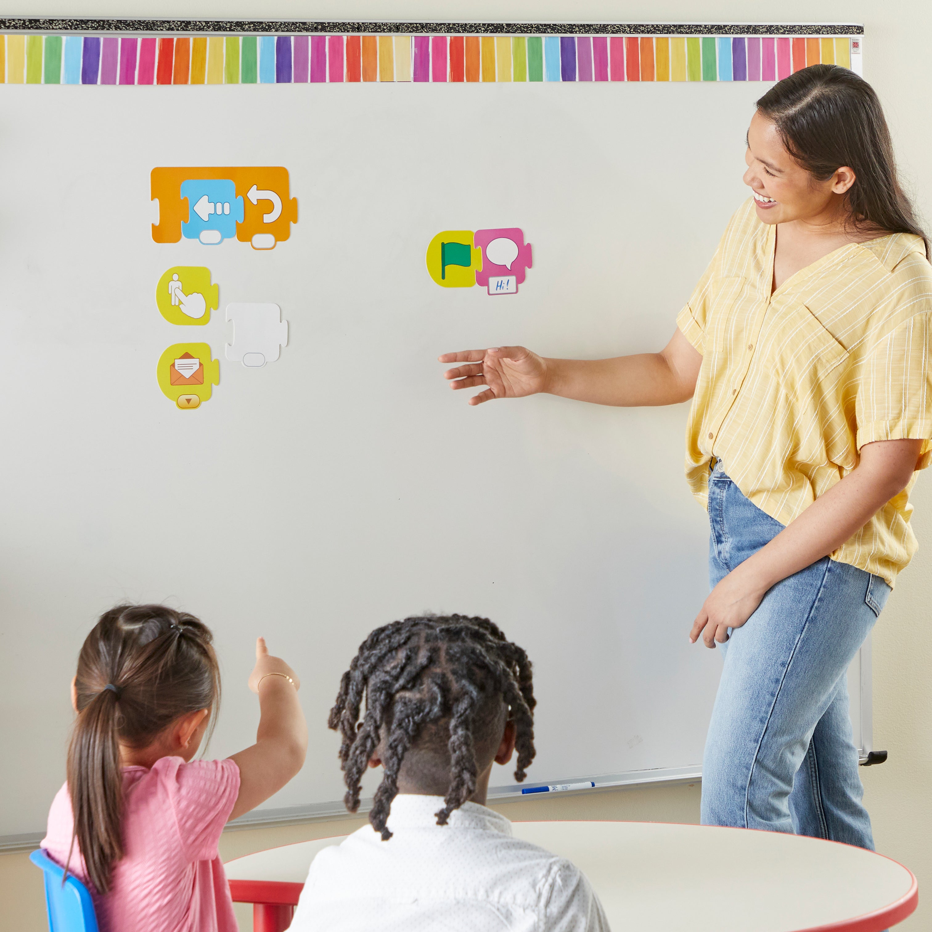 An educator at a whiteboard uses large magnetic ScratchJr blocks to demonstrate coding sequences. Two students watch and point as colourful icon-based tiles are arranged to show "unplugged" programming.