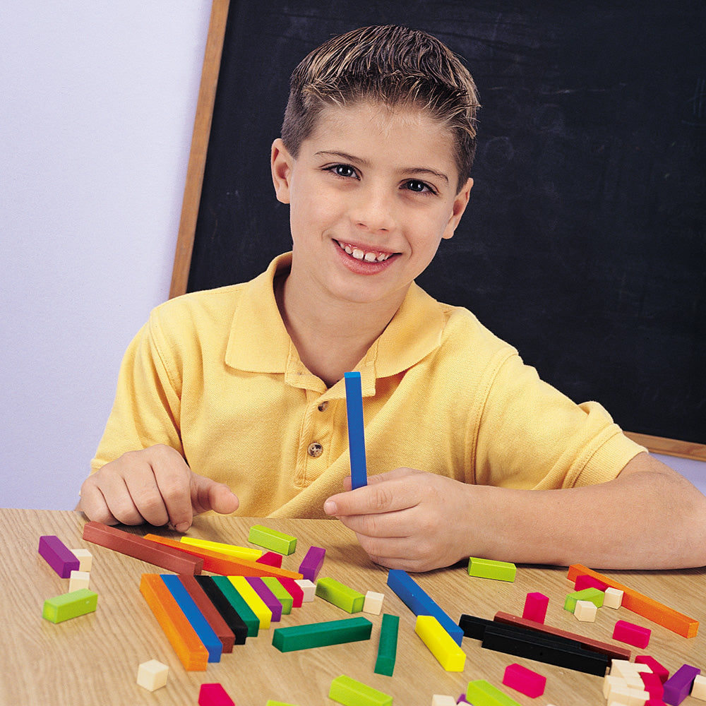 A young student smiling while holding a blue Cuisenaire Rod at a classroom desk. Various colourful plastic rods of different lengths are scattered across the table.