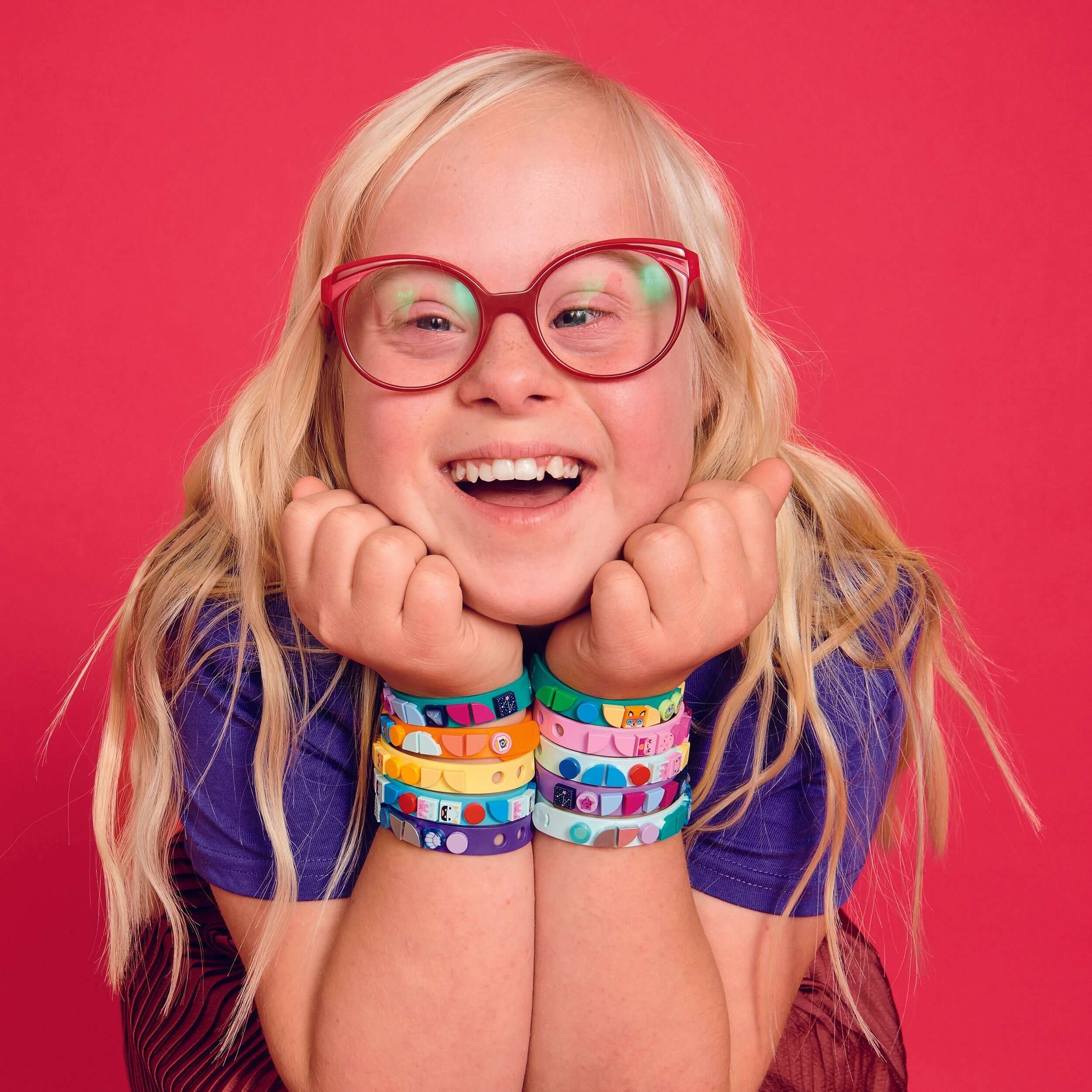 Child with colorful LEGO® DOTS starlight bracelets, smiling joyfully against a vibrant pink background.