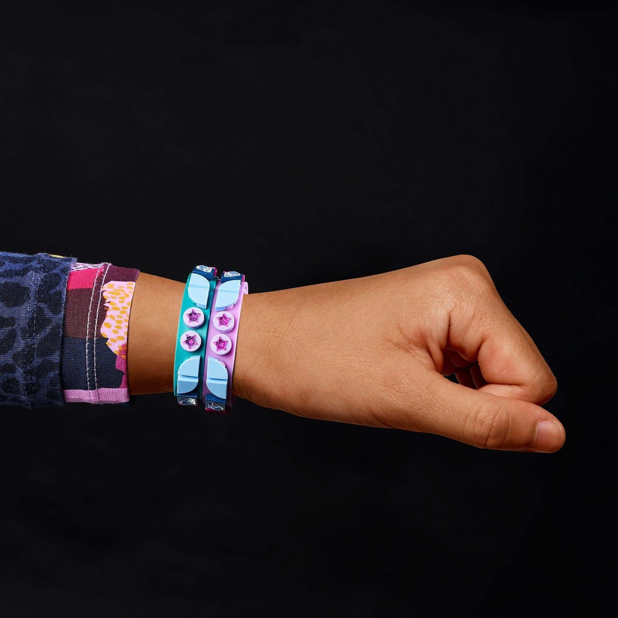 Child’s hand wearing LEGO® DOTS Starlight Bracelets in teal and purple with star tiles against a black background.