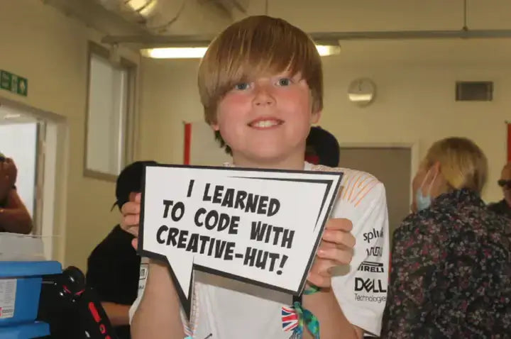 Young boy proudly holding a sign that says 'I learned to code with Creative-Hut' in a classroom setting.