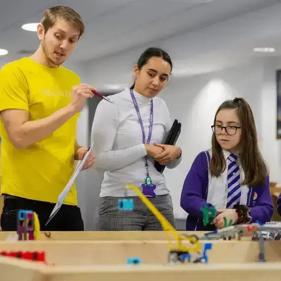 A man explains a robotics project to two girls, fostering STEM education and teamwork in a classroom setting.