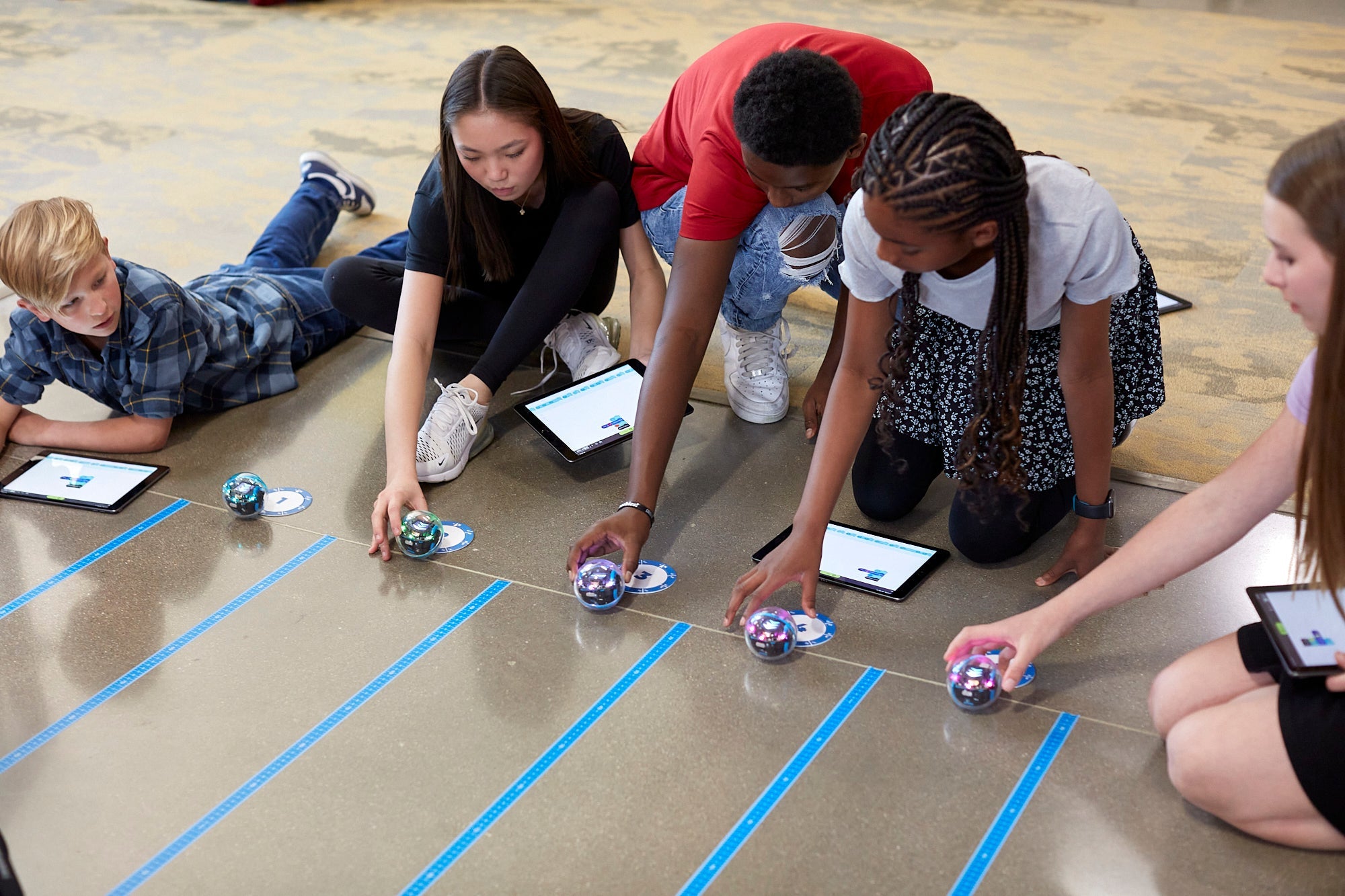Students engaged in coding with Sphero BOLT+ robots and tablets on the floor at a learning activity.