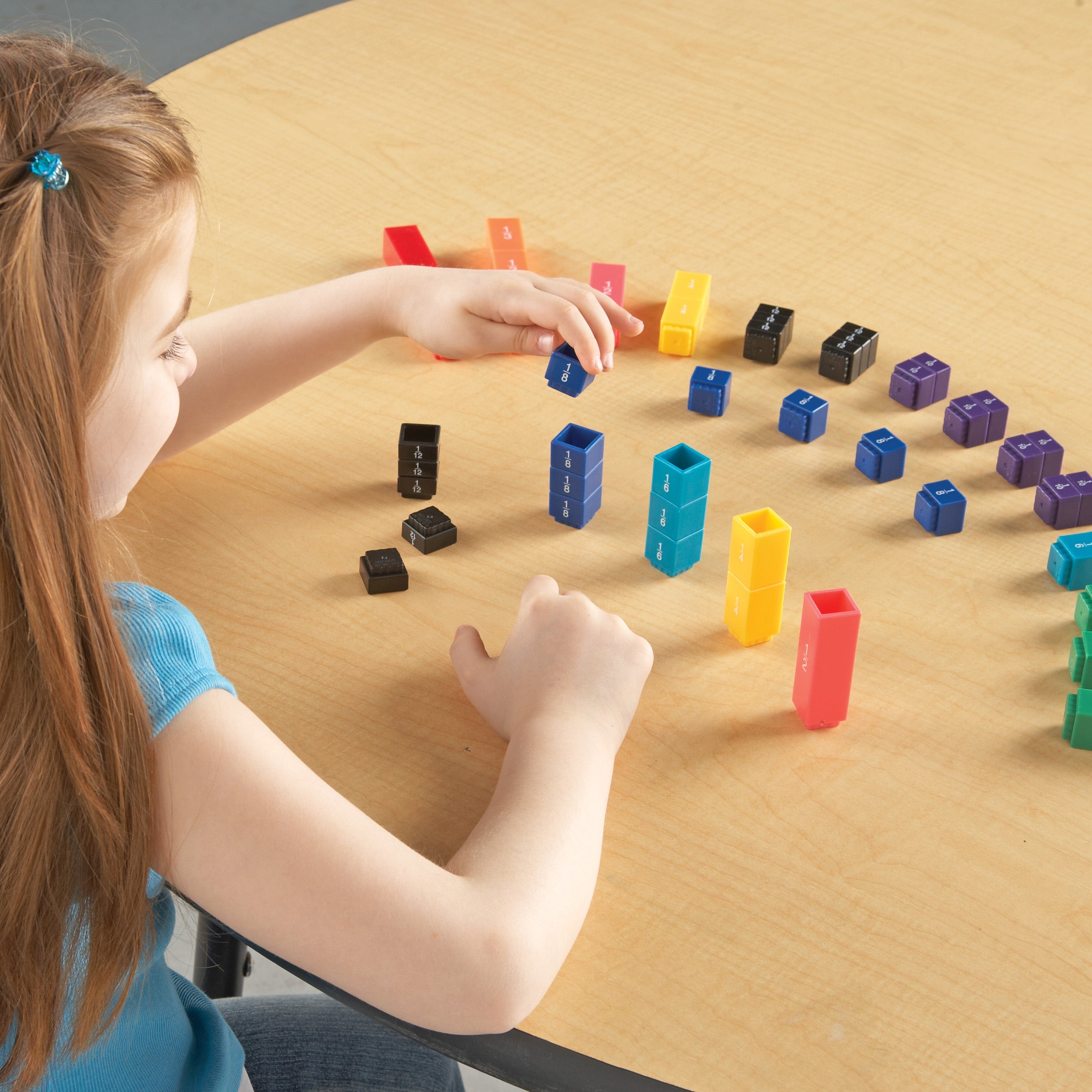 A high-angle shot showing a student at a wooden desk using the Fraction Tower® Cubes to explore mathematical concepts. The student is stacking blue cubes labelled "1/8" to compare their height with other colourful, interlocking fraction towers in red, yellow, and teal. Various other cubes representing different fractions are arranged across the desk, demonstrating how the set helps children aged 6+ visualise and compare fraction values and equivalence through hands-on play.