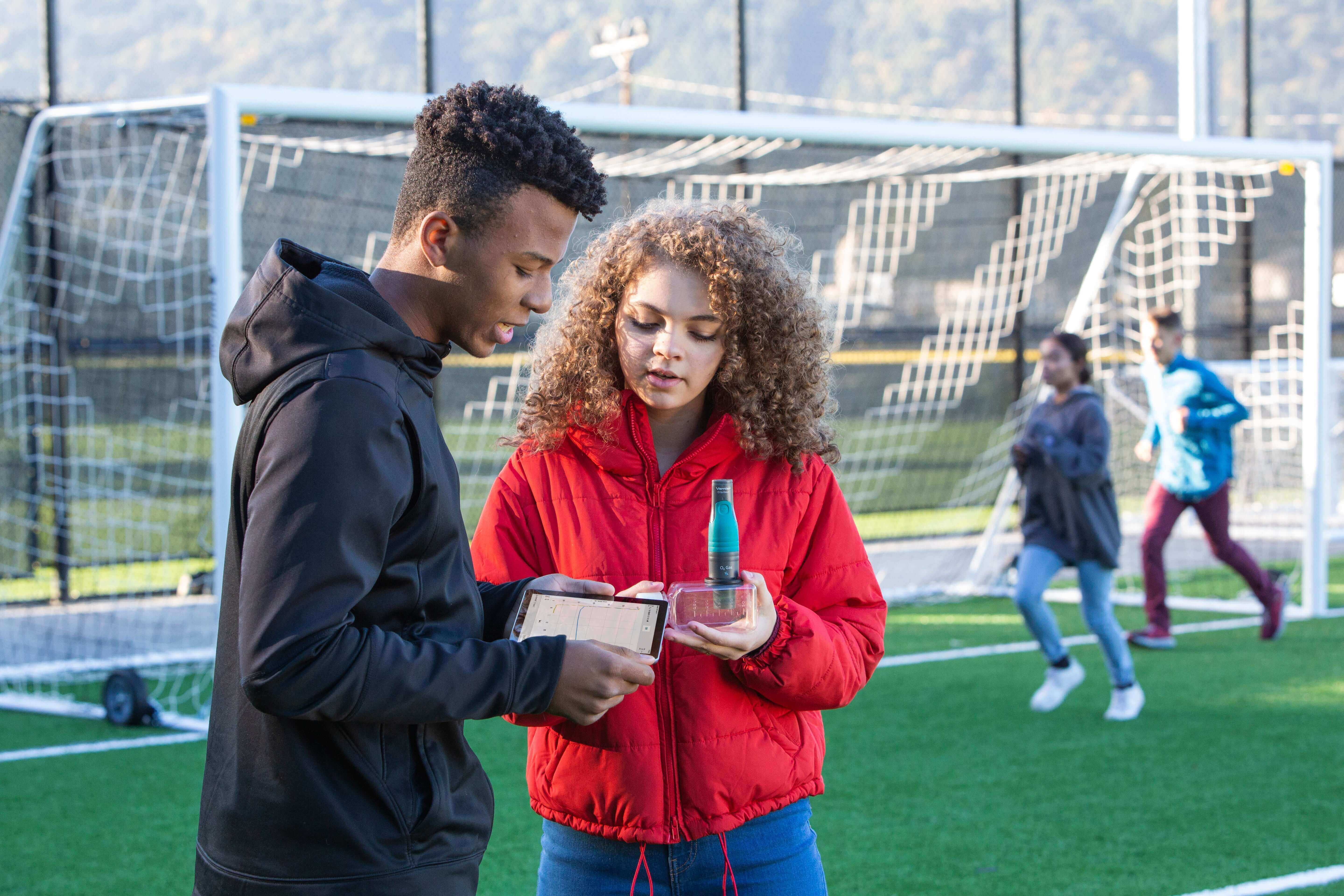 Two teens discussing a project on a soccer field, with goalposts in the background and their peers practicing.