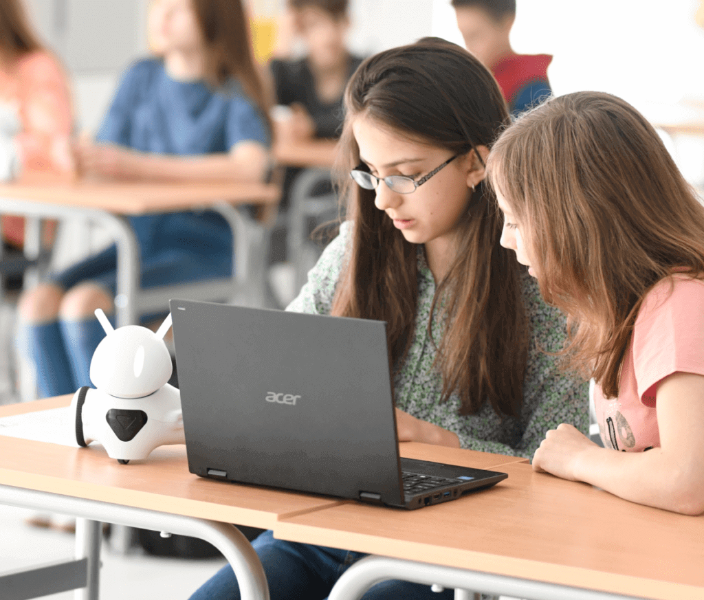 Two students collaborating with a laptop and a small robot in a classroom setting.