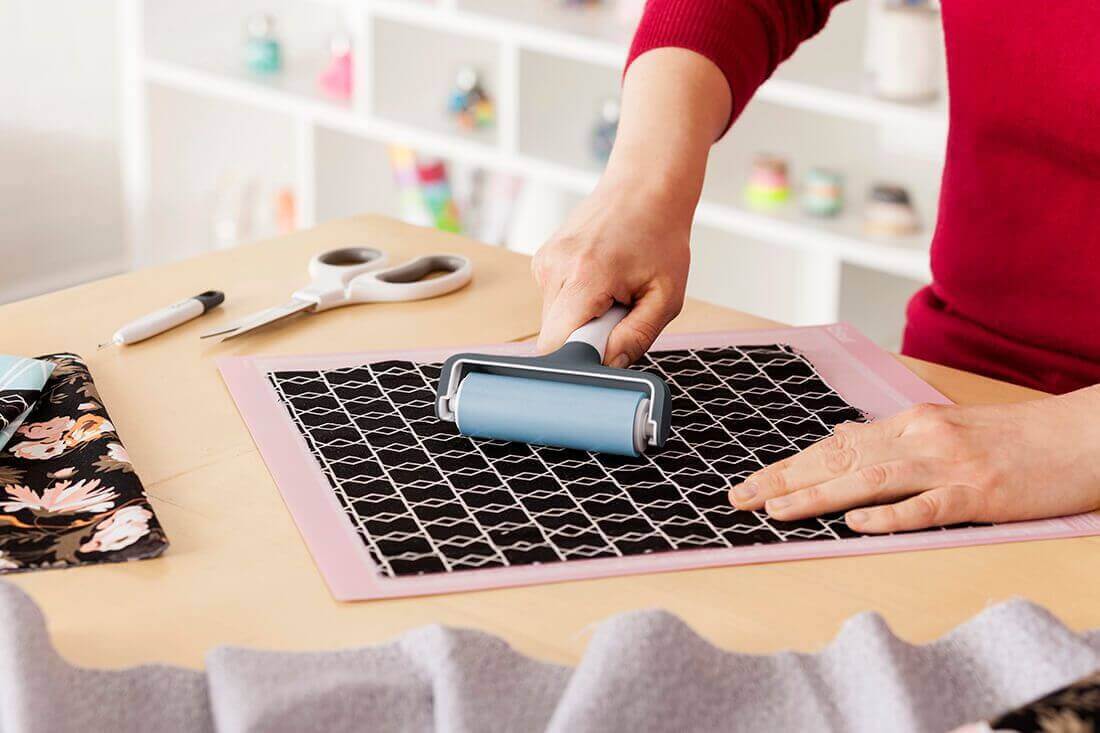 Person using a Cricut Brayer to apply fabric smoothly on a cutting mat in a crafting workspace.