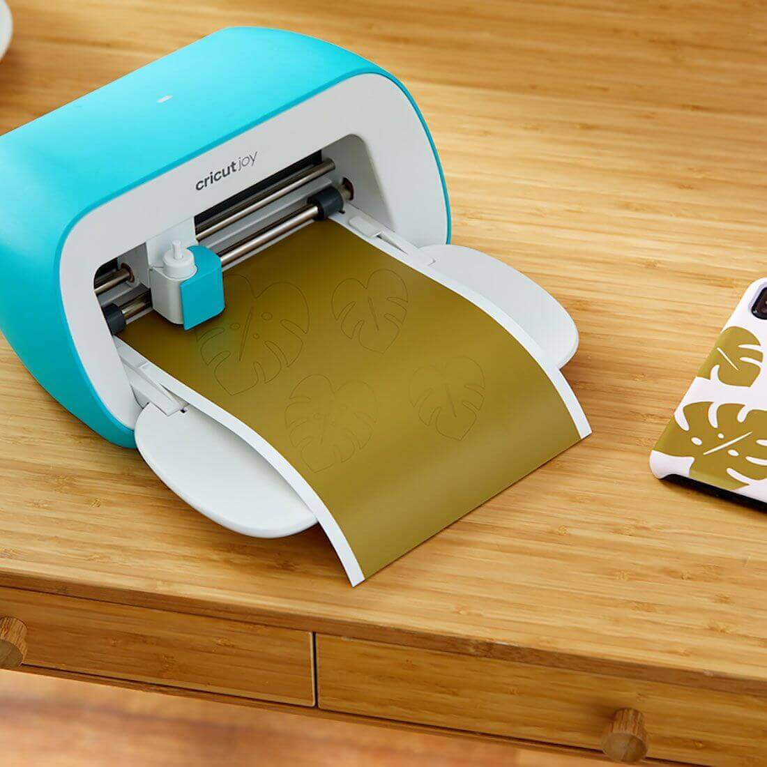 Cricut Joy cutting machine in use with a sheet of green Smart Vinyl on a wooden surface.