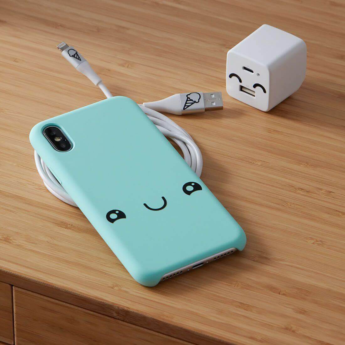 Cute mint phone case with smiley face, USB cable and cheerful charger block on wooden surface.