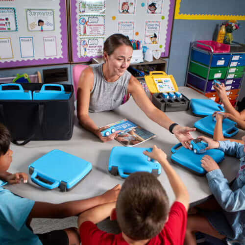A teacher engages with students using educational tools at a classroom table, promoting interactive learning.