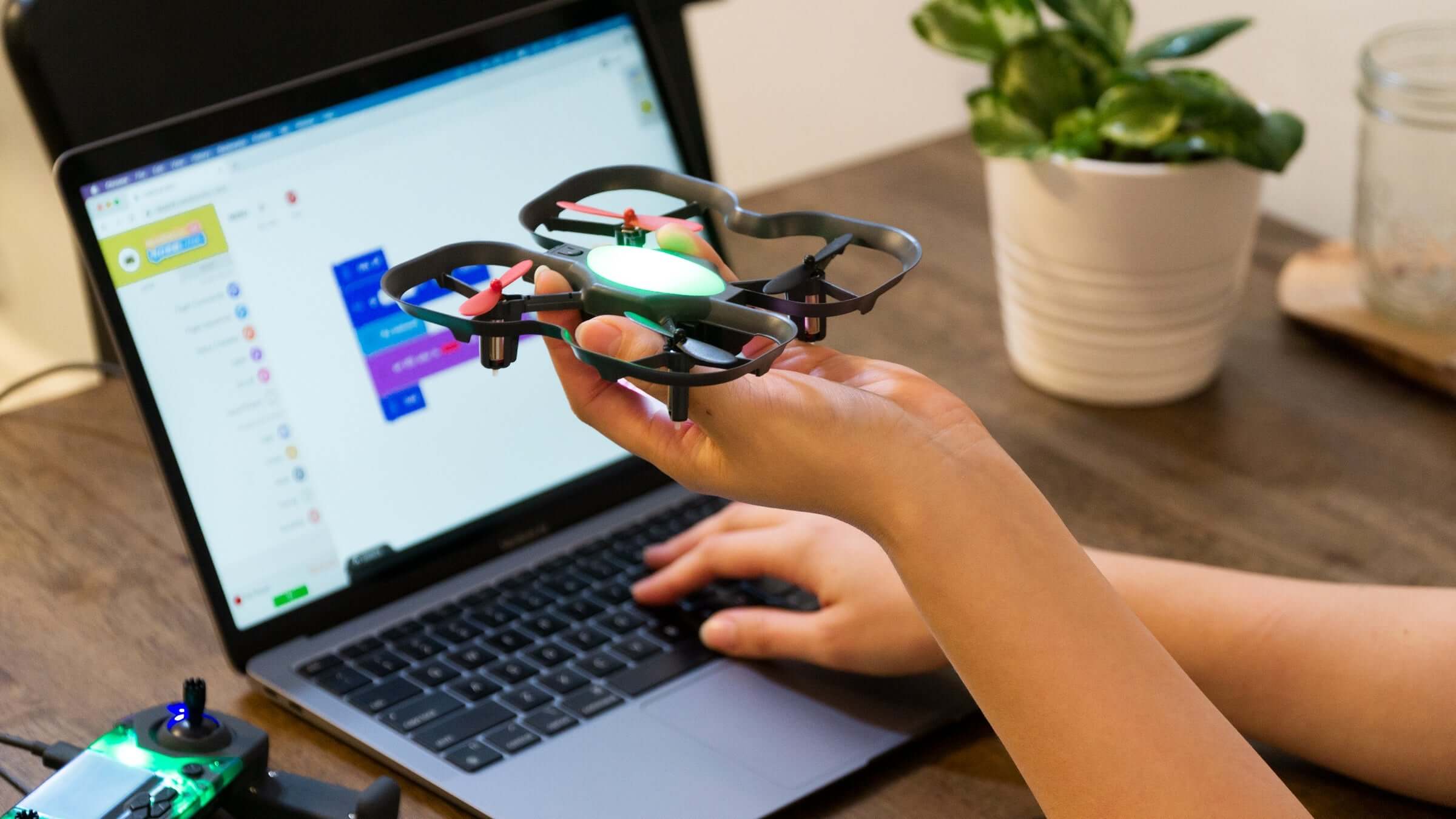 Person controlling a drone while using a laptop with coding software on the screen.