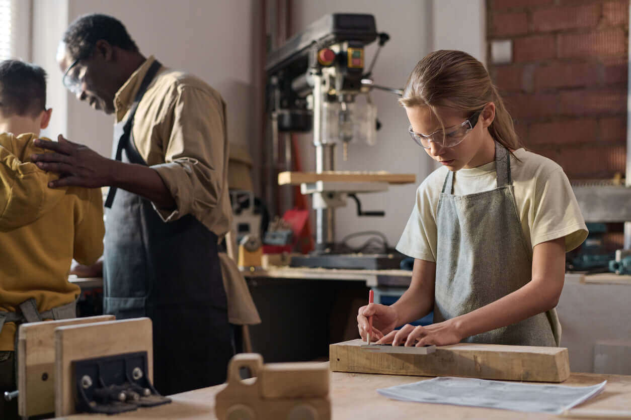Girl in workshop using tools while a man assists a boy in woodworking teaching environment.