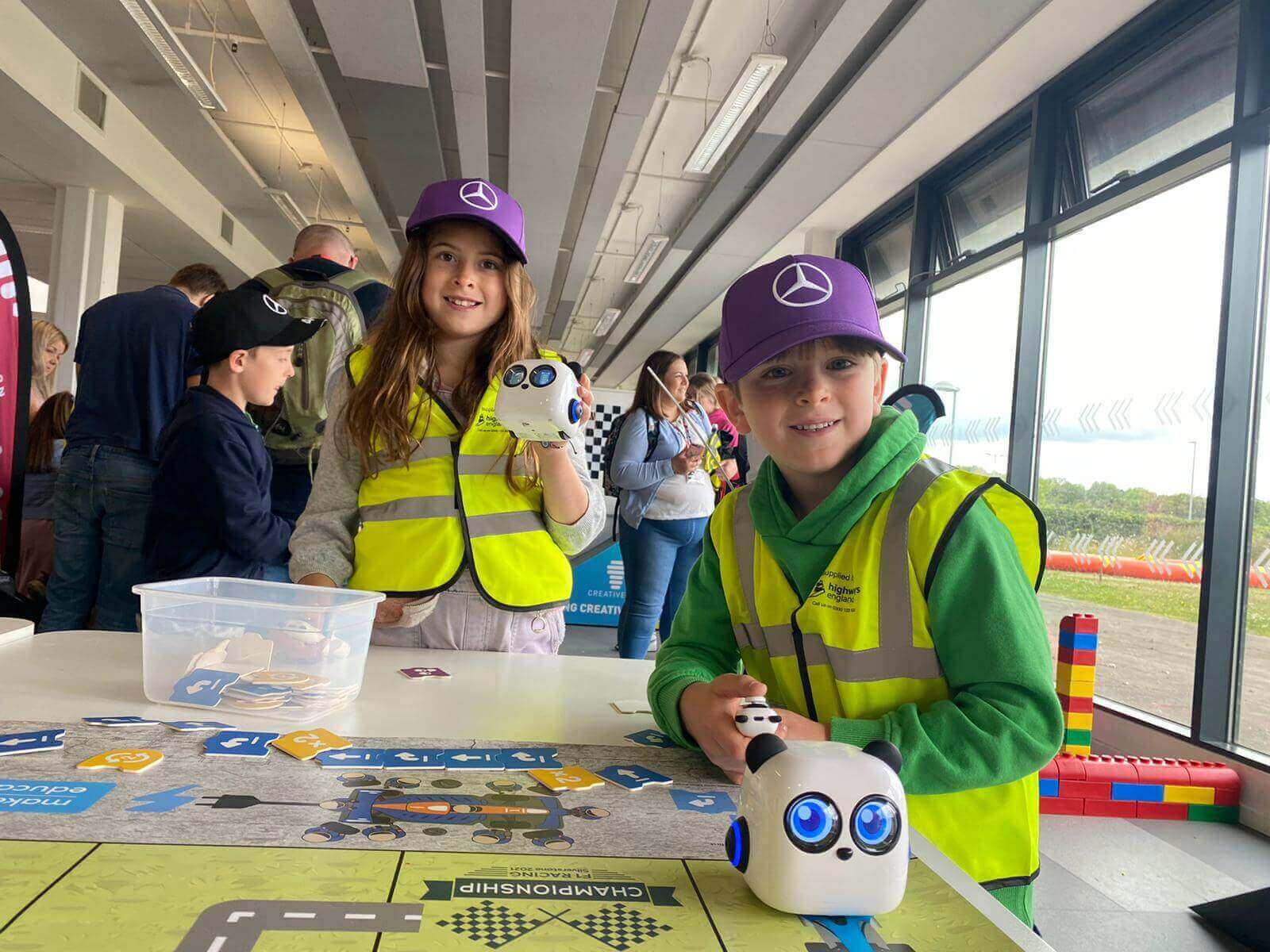 Two children in safety vests and caps interact with robotic toys at a hands-on learning event indoors.