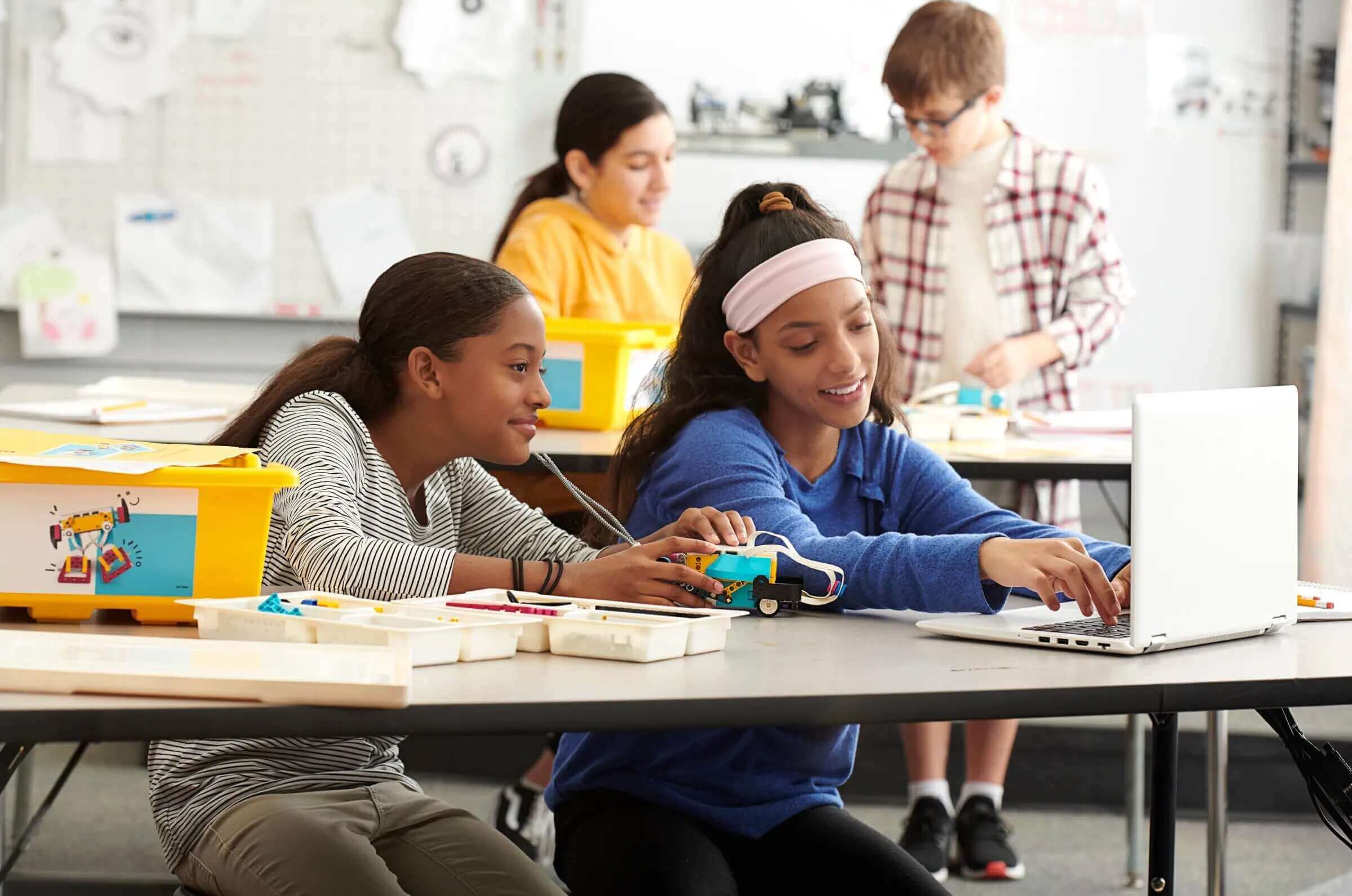 Two girls collaborate on a robotics project at a table, using LEGO materials and a laptop in a classroom setting.