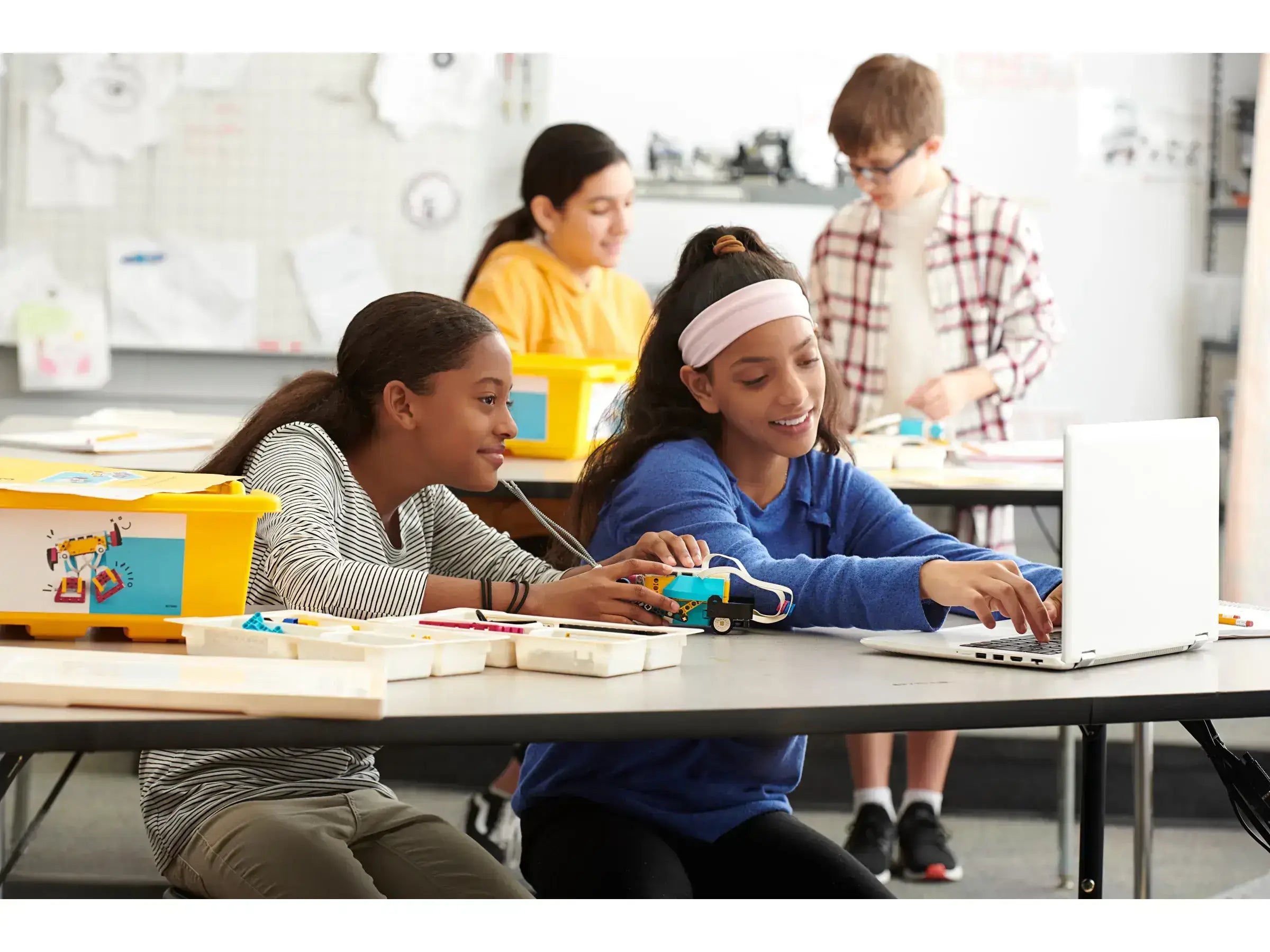 Two girls collaborating on a robotics project at a classroom table, with others working in the background.