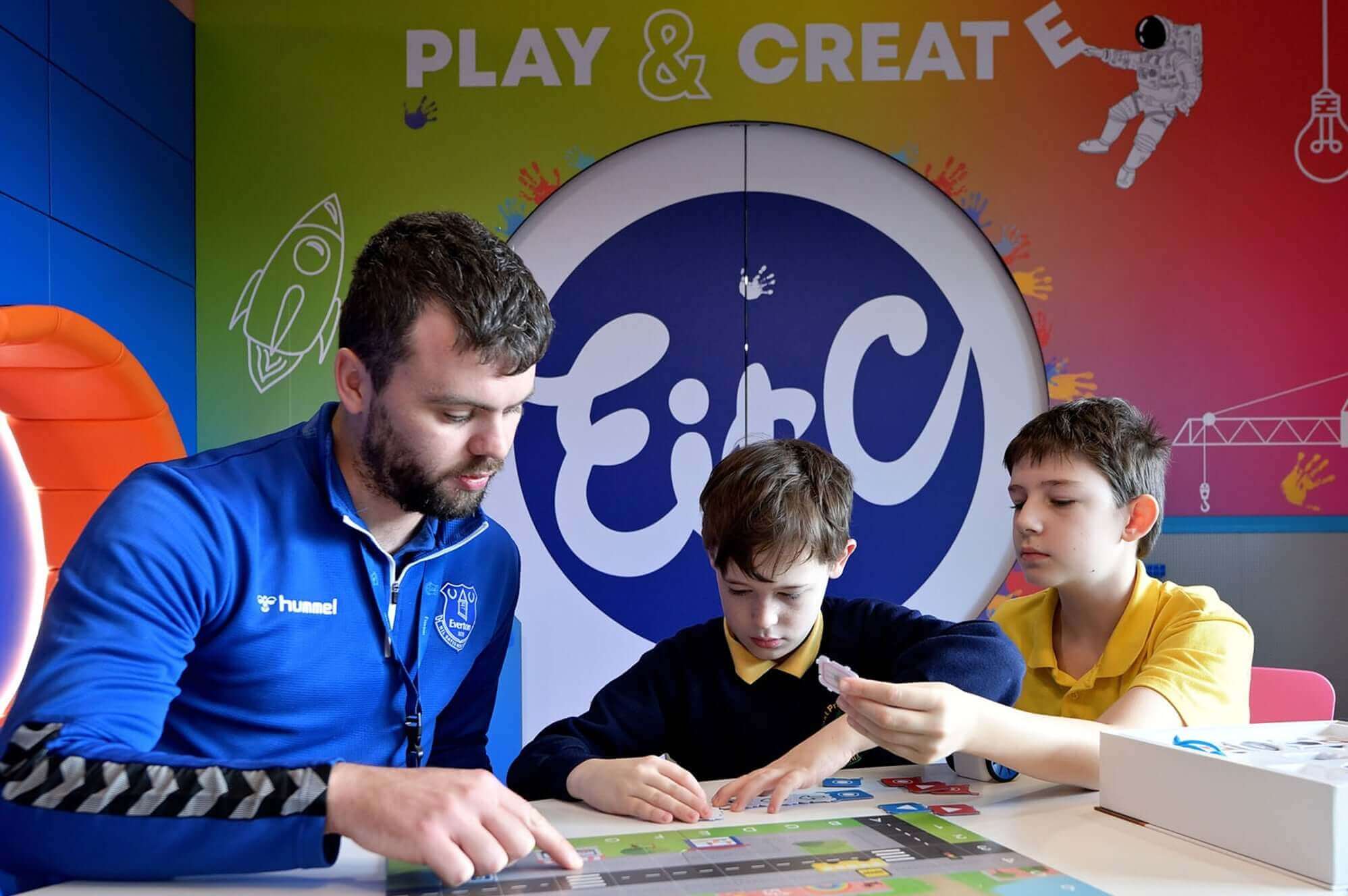 A man teaches two boys while playing a game in a colorful educational environment with the text 'Play & Create'.