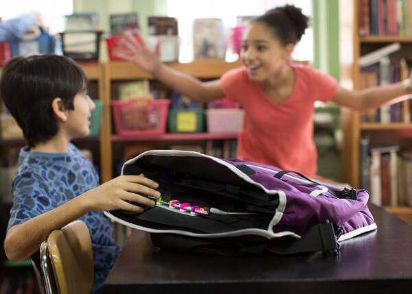Two kids interacting at a classroom table, one excitedly reaching towards a backpack filled with littleBits STEAM Student Set tools.