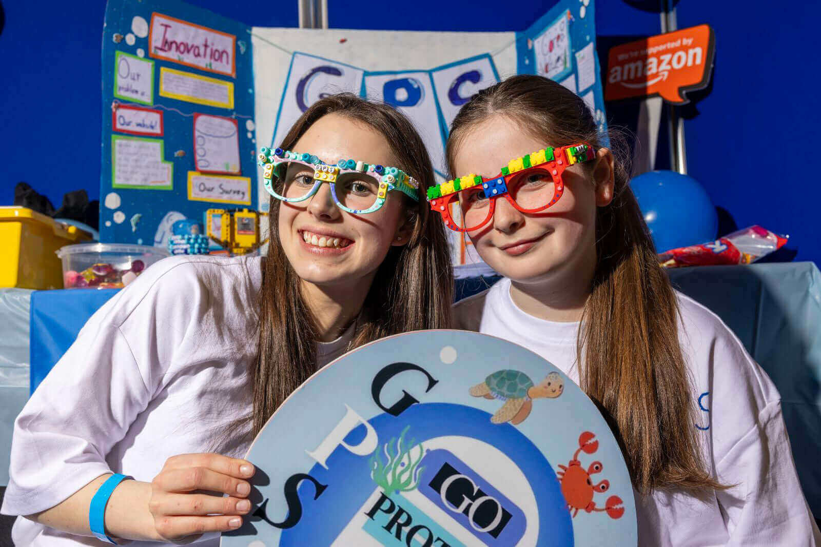 Two girls with creative glasses pose proudly with their project display at a school science fair.