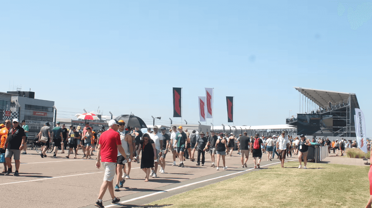 Crowd of people walking at a motorsport event under a clear blue sky.