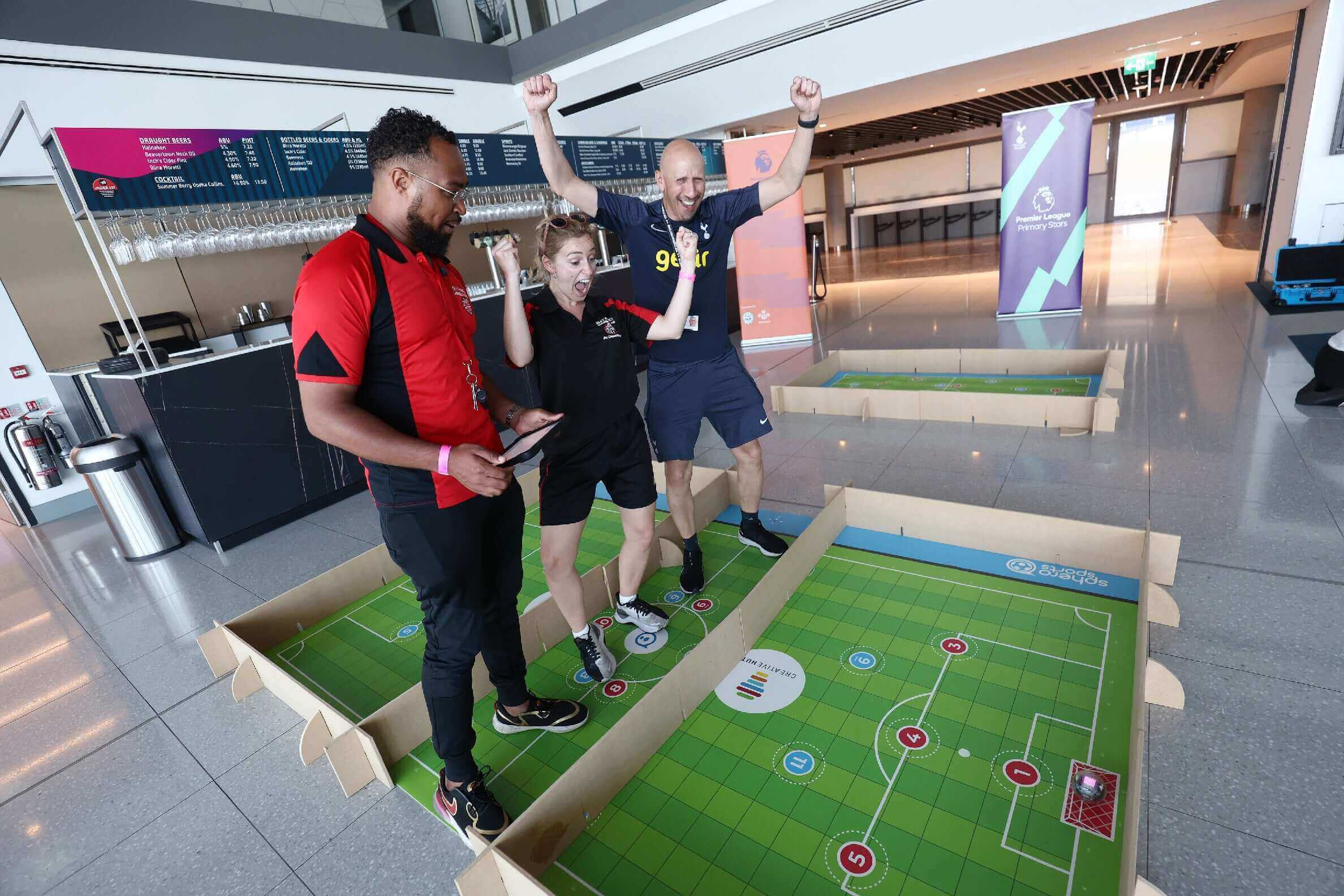 Three excited participants celebrating while engaging in a team-building game on a soccer-themed board in a brightly lit venue.