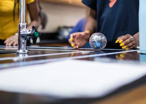 Person with yellow nails rolling a robot toy on a lab table, showcasing hands-on learning in computer science.