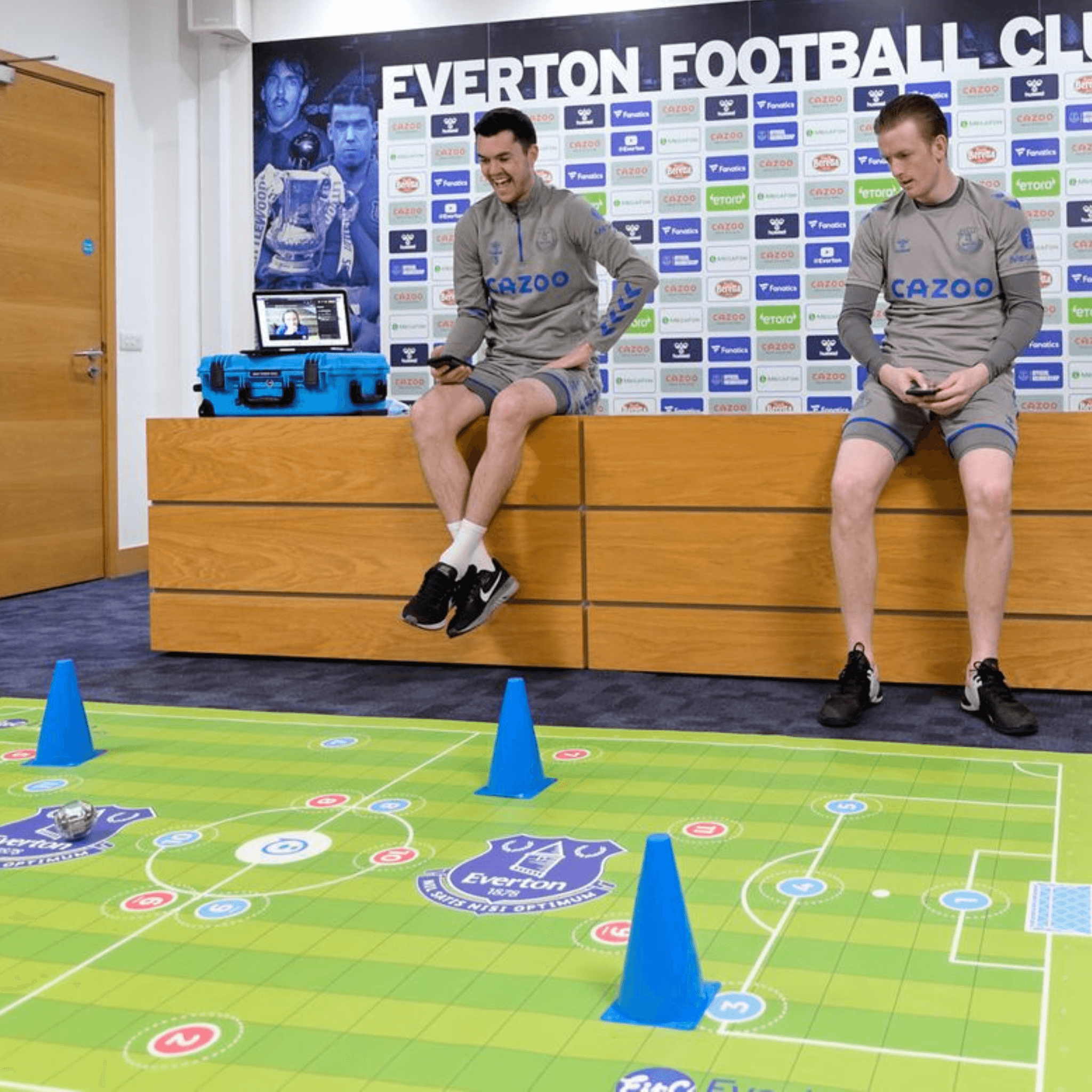 Two athletes seated on a bench with an Everton Football Club backdrop, interacting with a coding sports mat.
