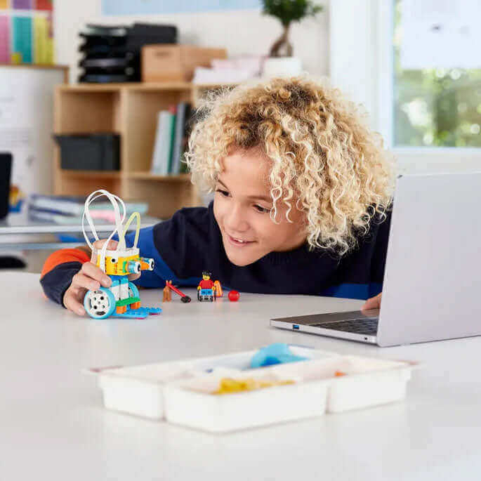 Child playing with a colorful robot toy at a table next to a laptop, fostering creativity and STEM skills.