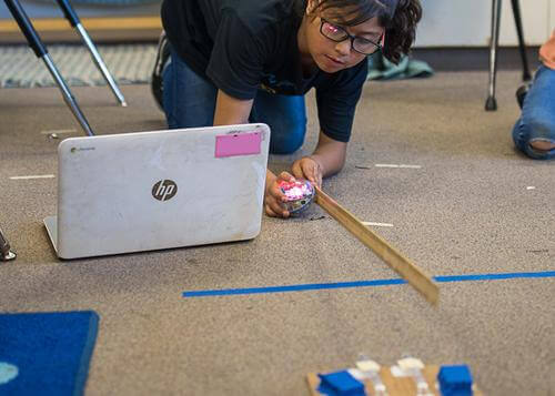 Student engages with Sphero robot while using a laptop to explore coding concepts in a classroom.
