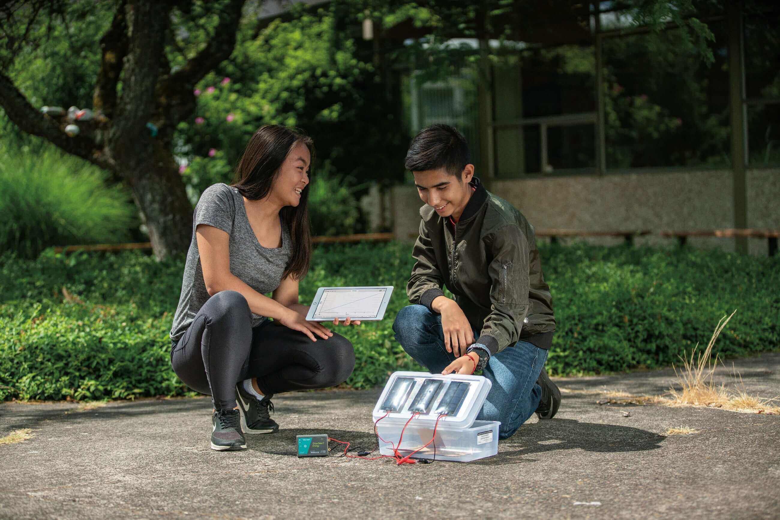Two students working on a project outdoors, using a tablet and a device with multiple components.