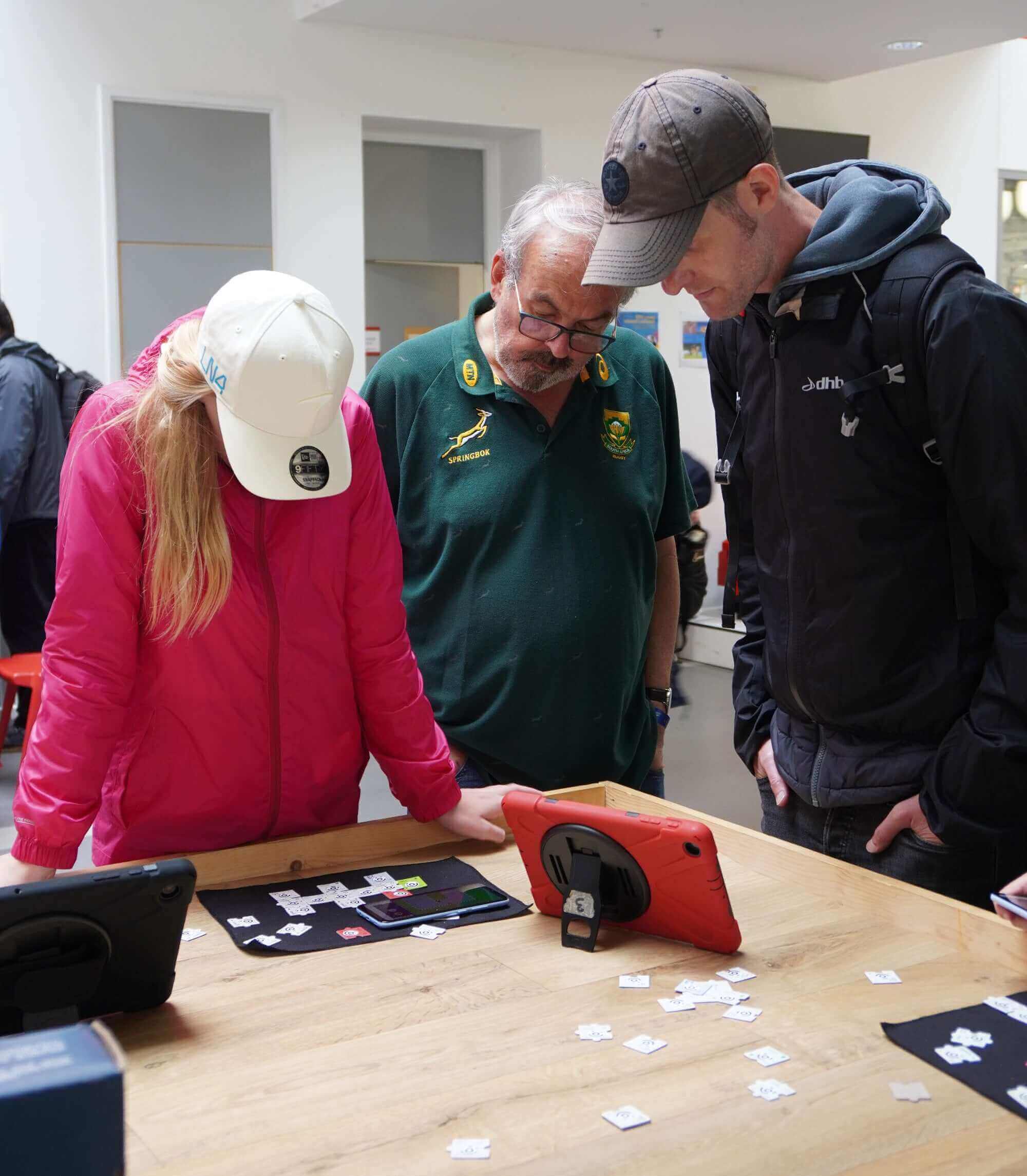 Three people engaged in a learning activity, using tablets at a table with scattered game pieces.