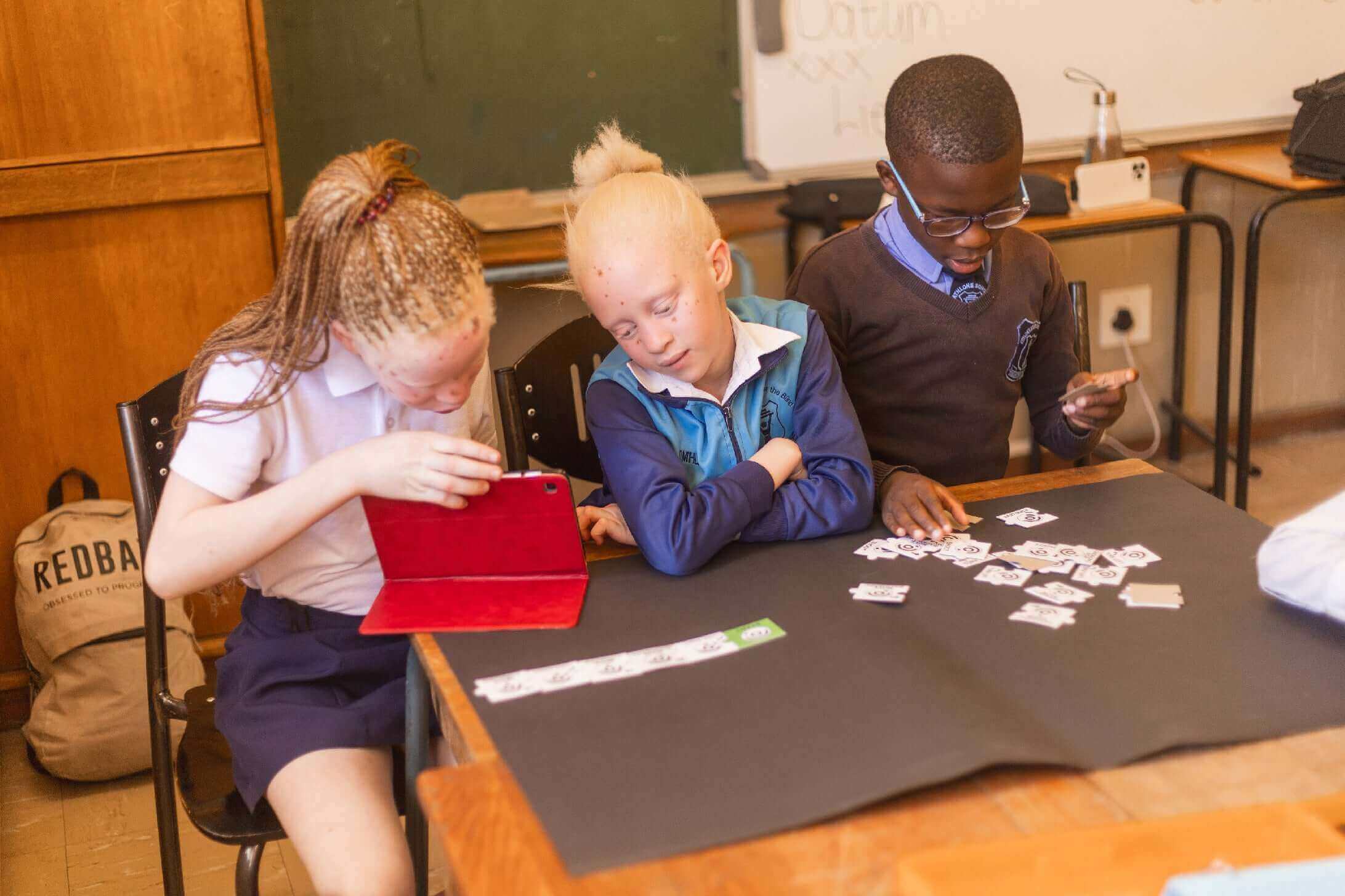 Children engaged in a learning activity at a classroom table, using a tablet and working with cards.